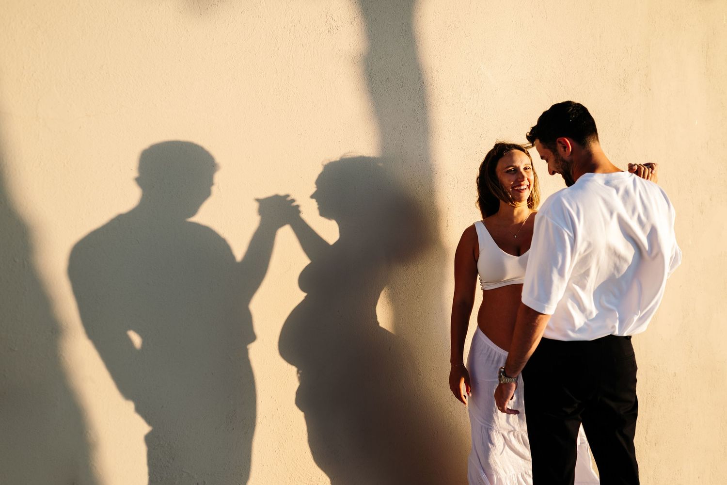 Silhouettes cast dramatic shadows against beige wall during intimate outdoor photoshoot.