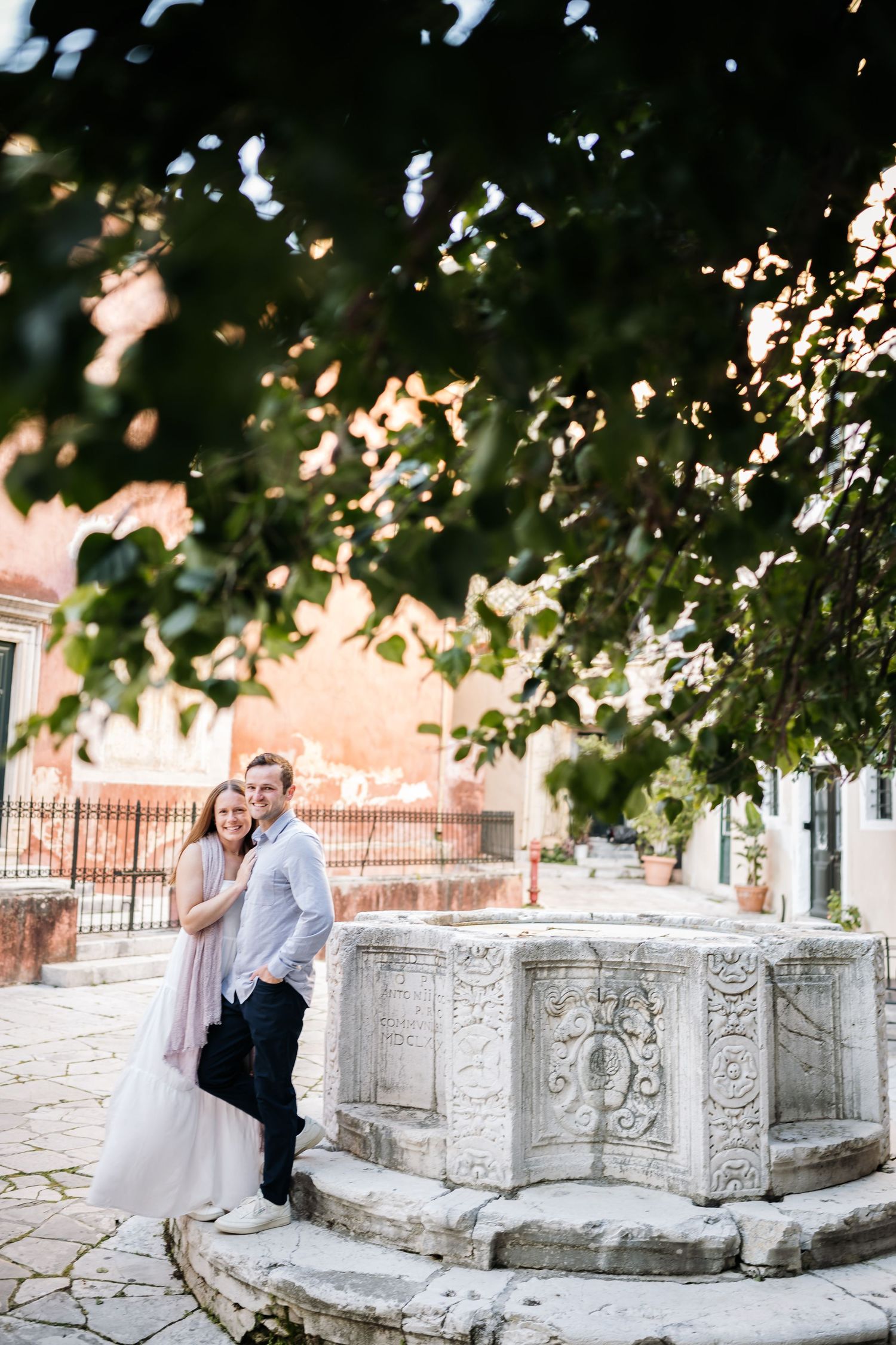 Couple sitting on ornate stone bench under leafy green trees.