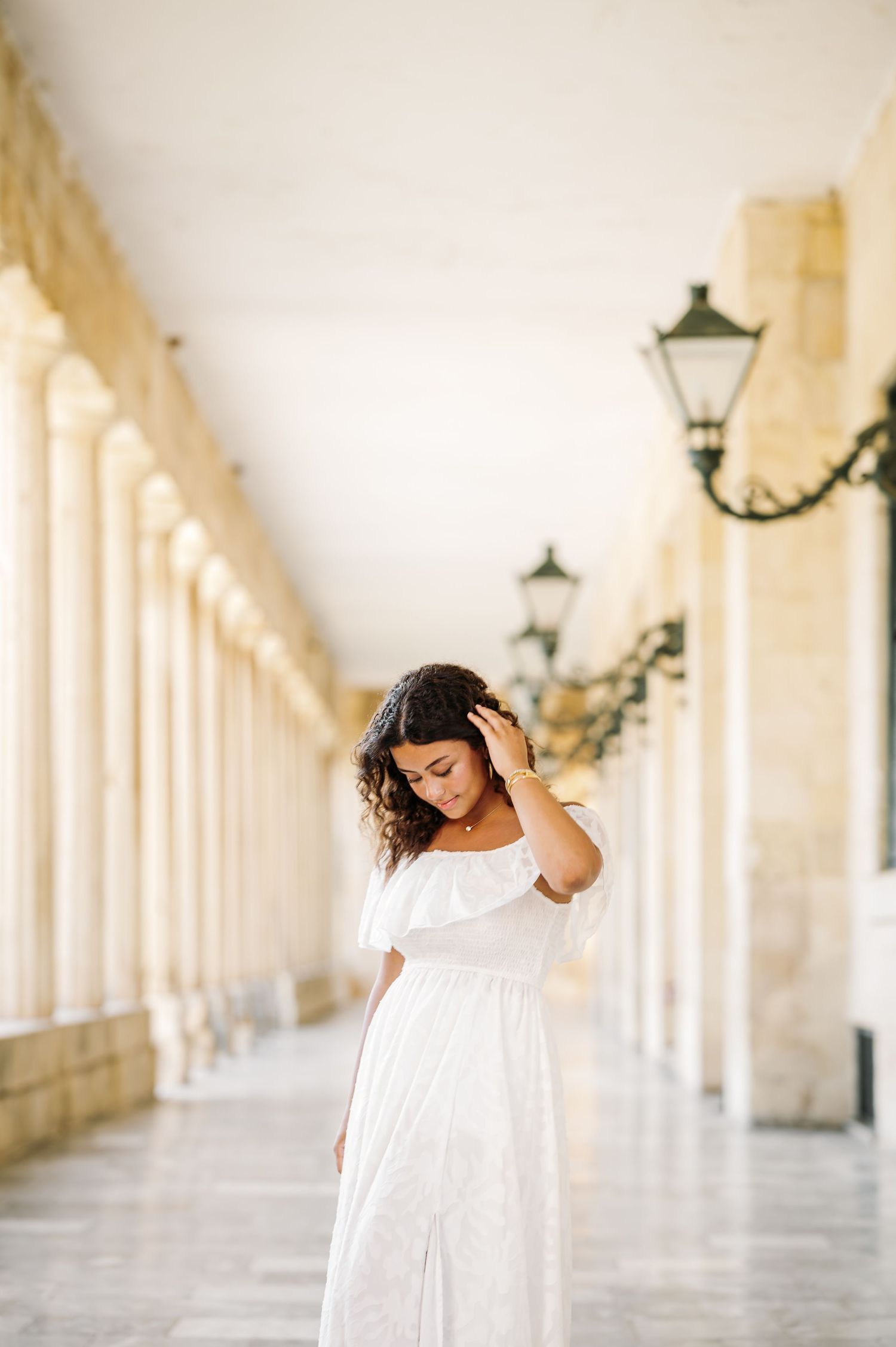 Person in white dress poses gracefully against classical columns in soft lighting.