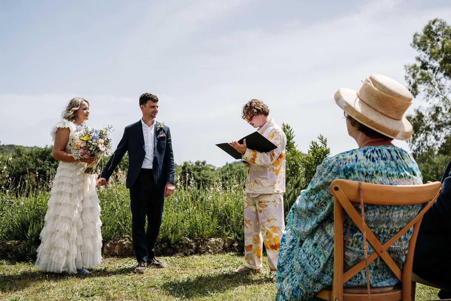 Intimate outdoor wedding ceremony with guests seated on wooden chairs in a natural setting.