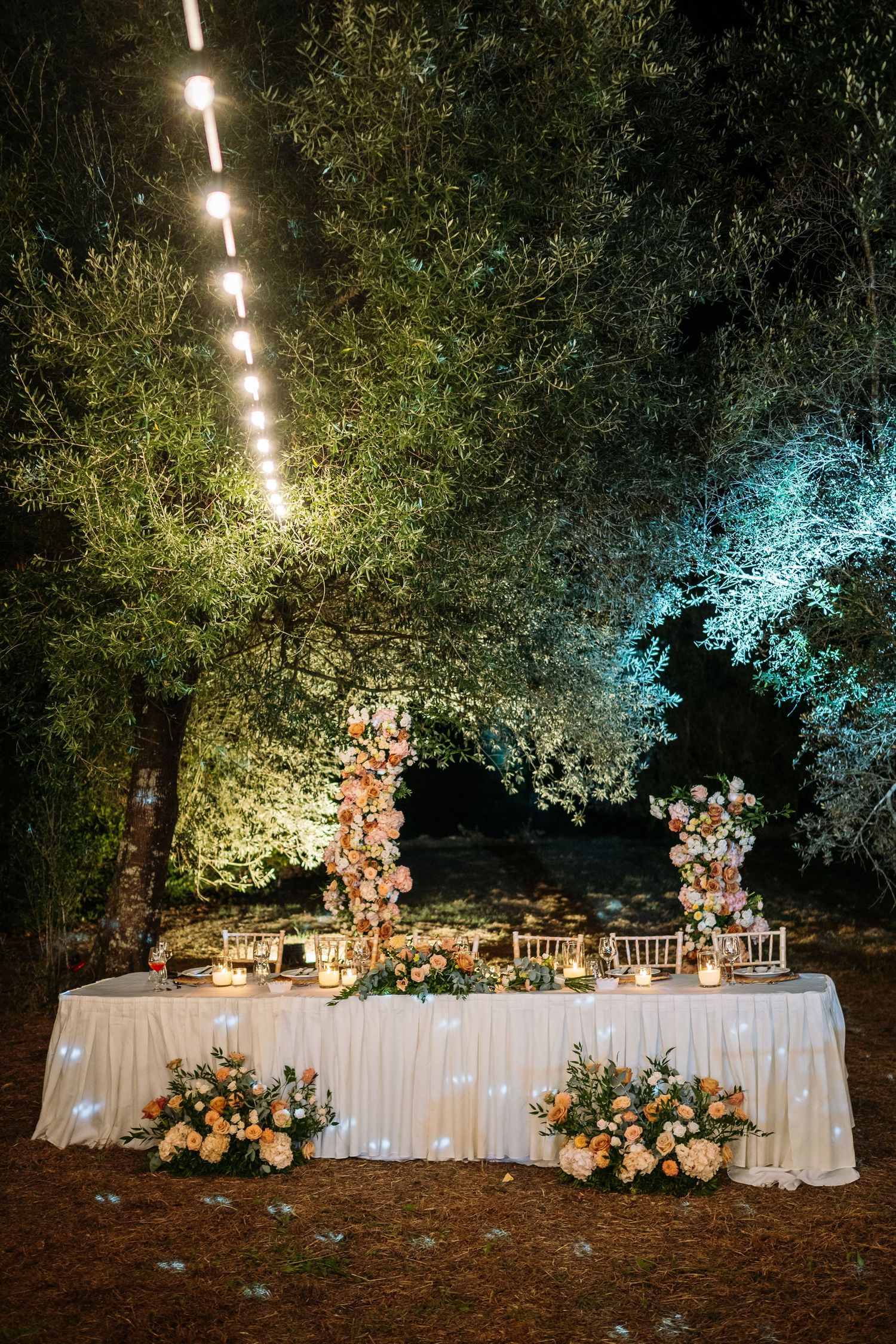 Elegant outdoor reception setup with string lights and floral arrangements on white-draped tables.