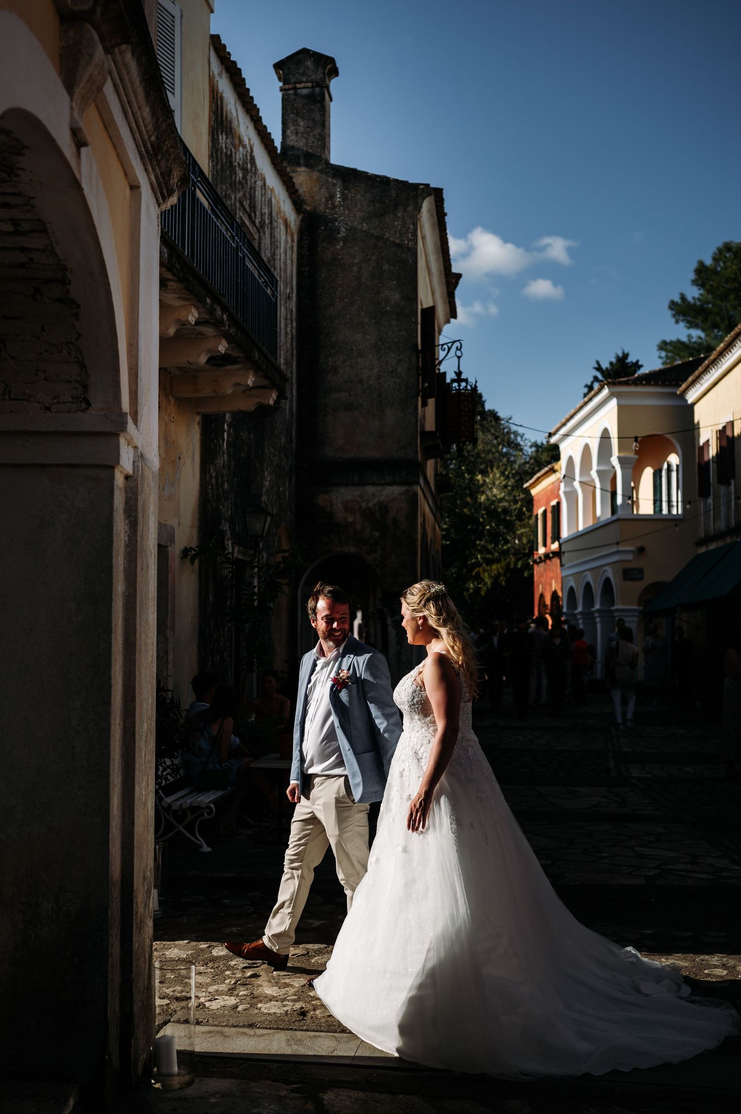 Wedding couple walking through a sunlit Mediterranean courtyard with stone architecture.