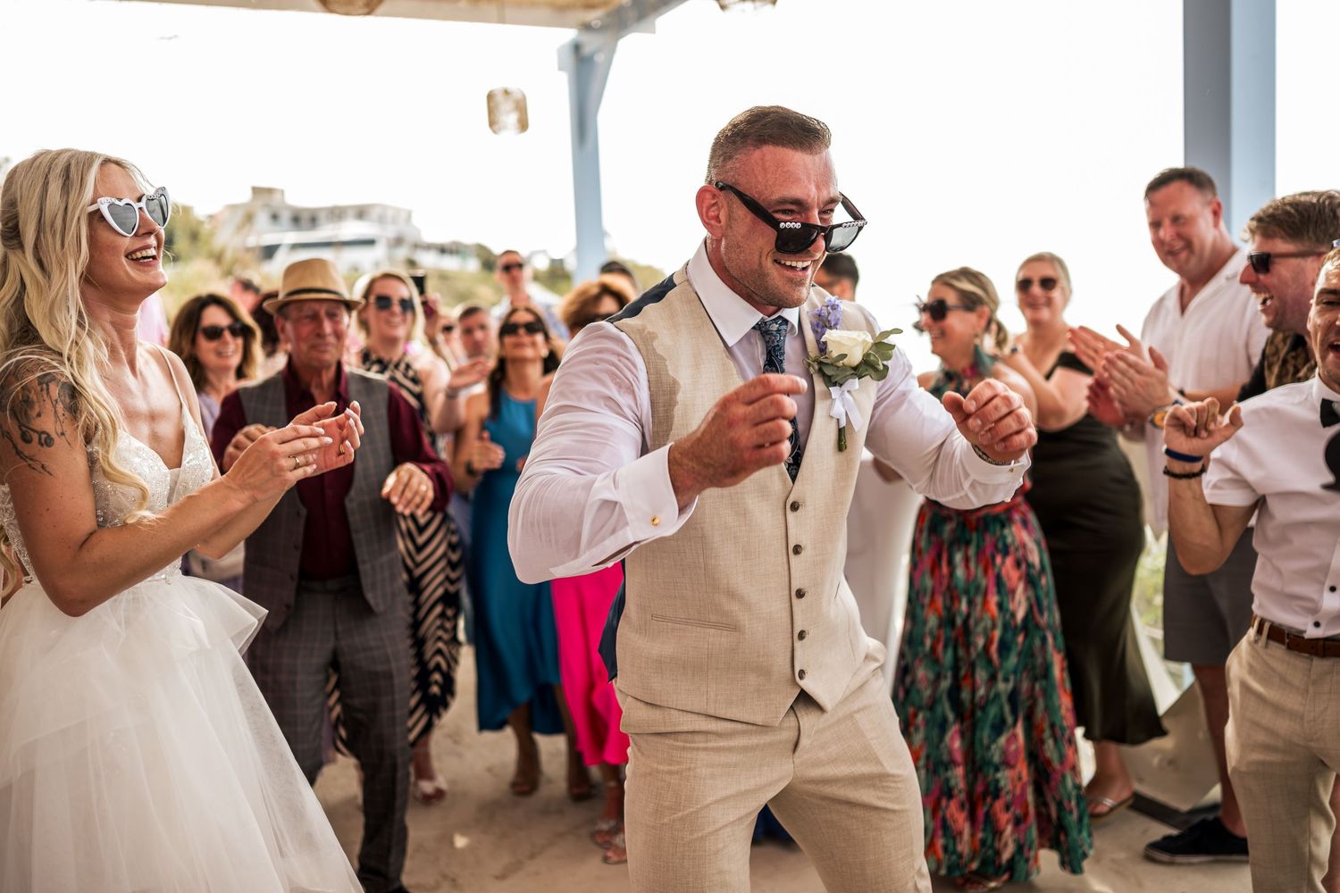 Wedding guests in summer attire dance and celebrate under a white covered pavilion by the beach.