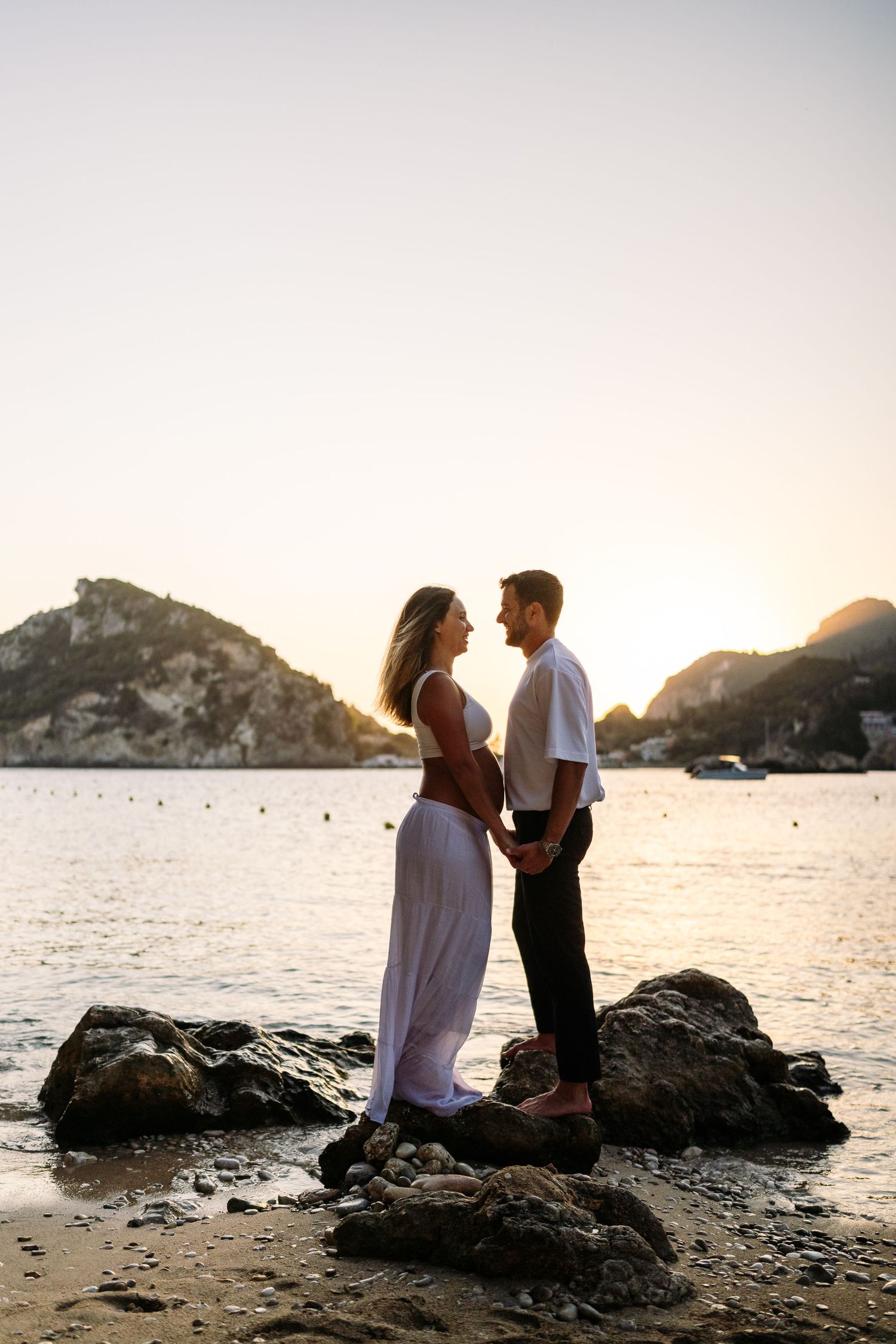 Couple shares intimate moment on rocky beach during sunset with coastal mountains in background.
