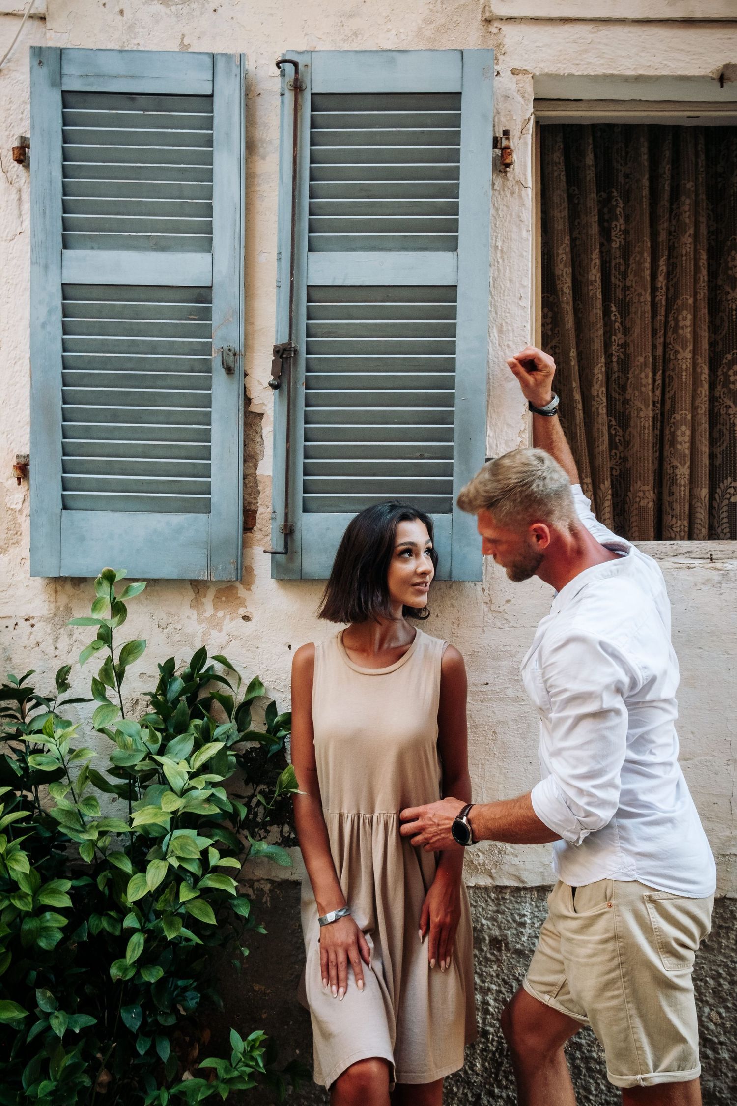 Couple posing playfully against wall with blue shutters and green foliage.