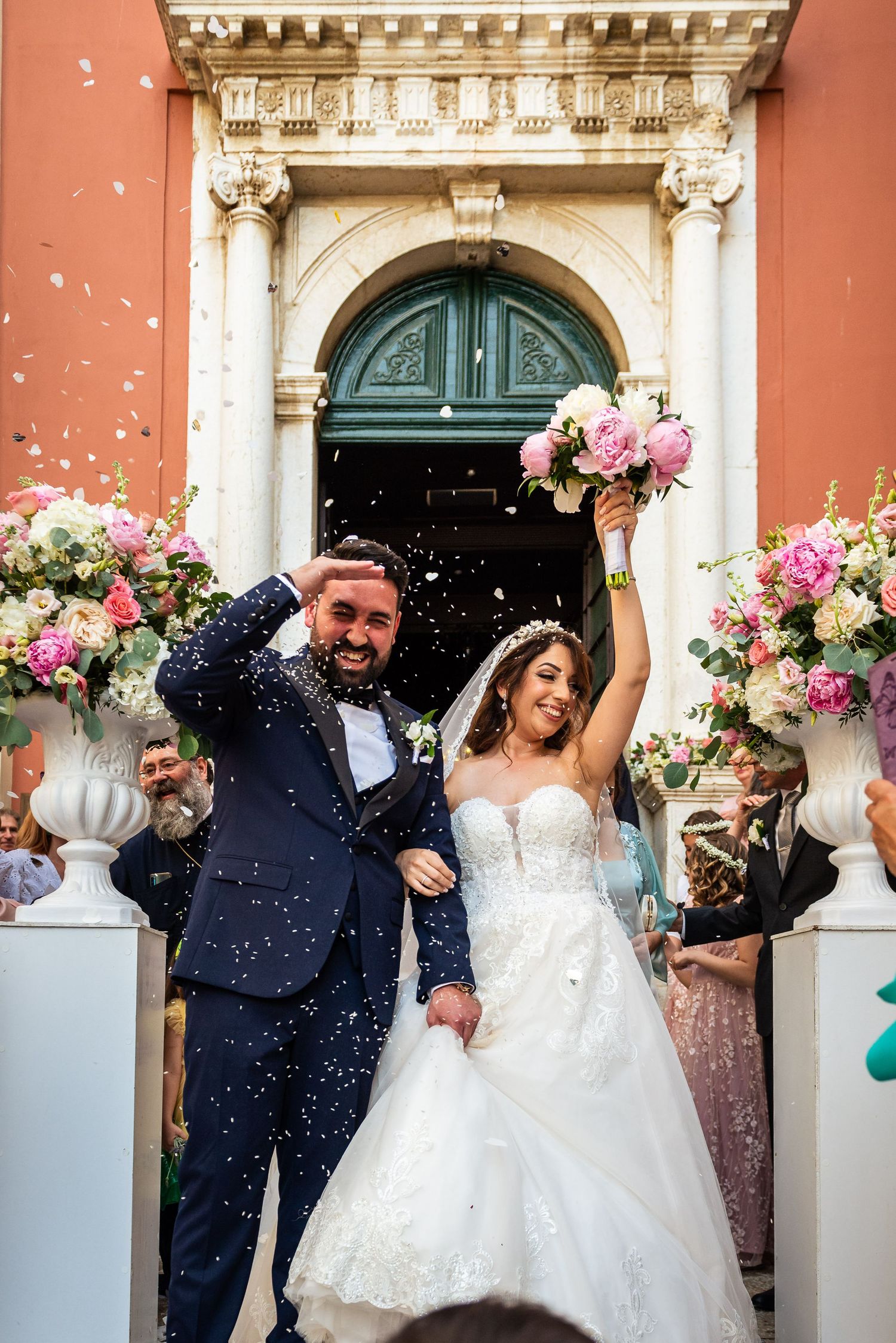 A joyful wedding celebration with confetti outside a coral-colored historic building.