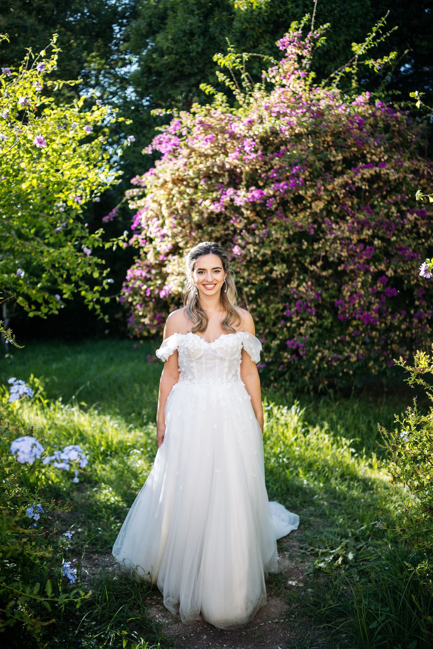 Bride in flowing white dress poses in garden setting with blooming pink flowers.