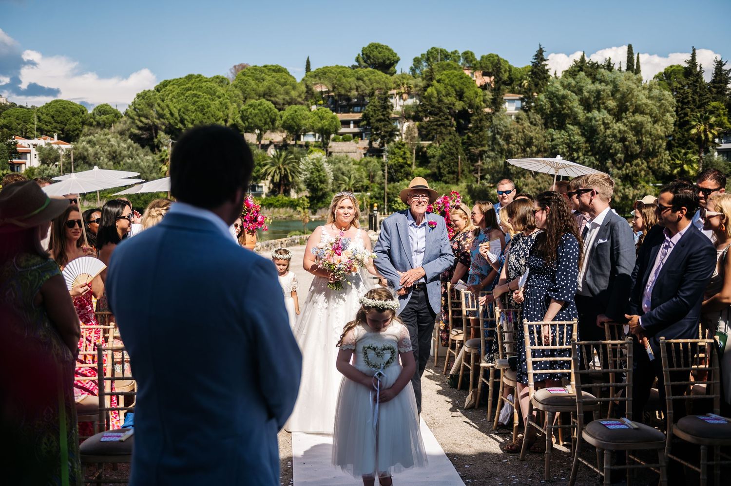 Wide view of outdoor wedding ceremony with guests seated in wooden chairs.