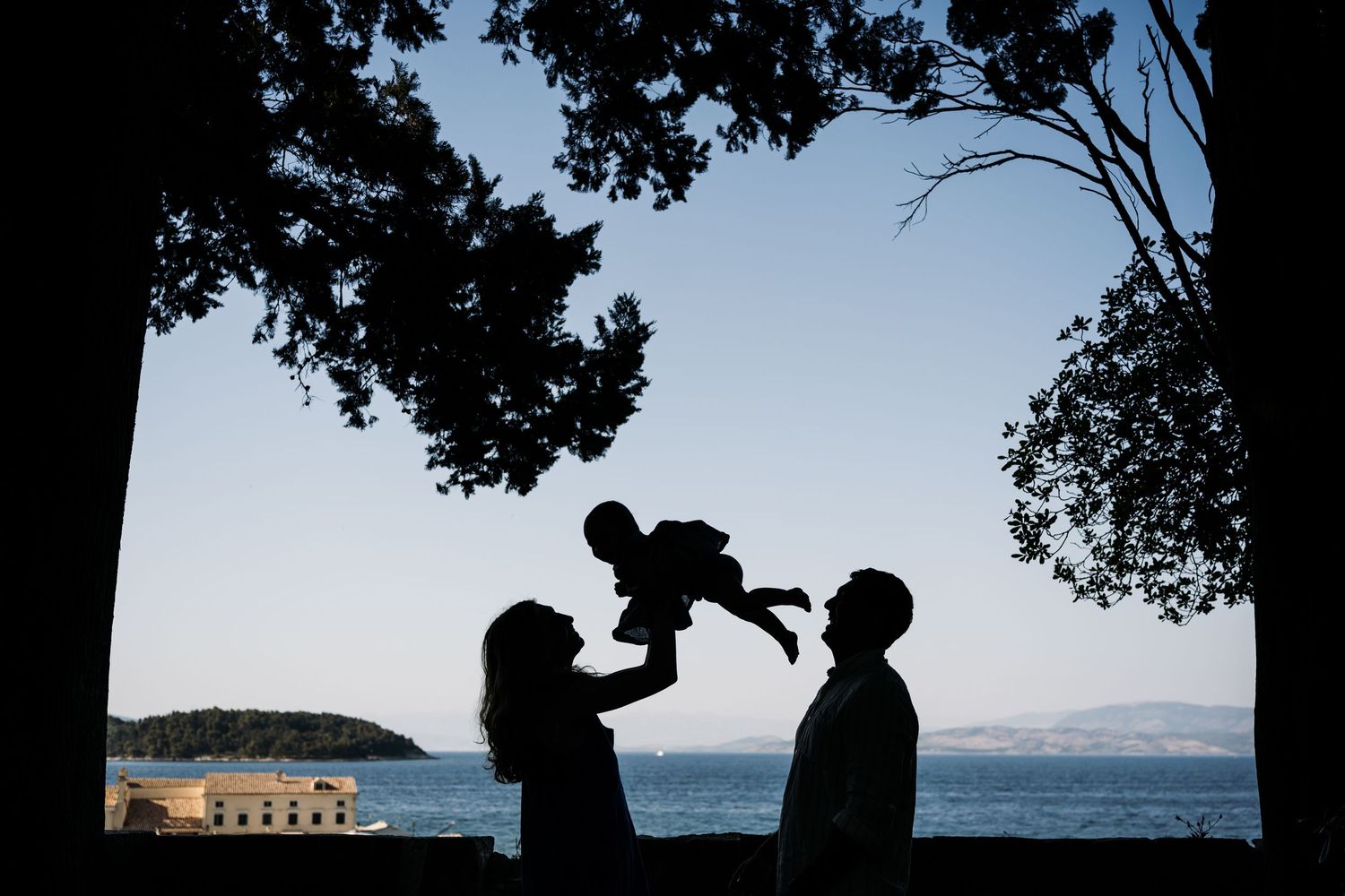 Silhouette of family playing with baby against ocean backdrop at sunset.
