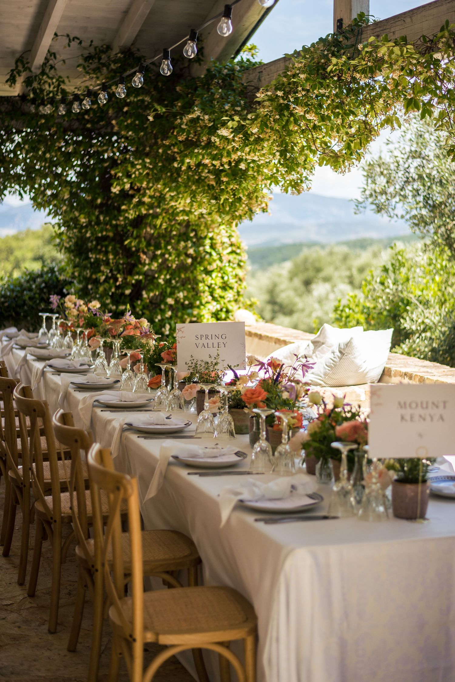 Outdoor wedding reception setup with white tablecloths and floral centerpieces under natural canopy.