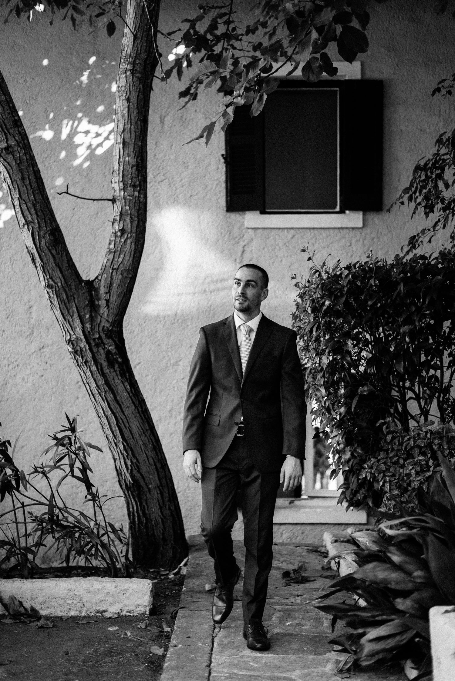 Black and white portrait of a person in a suit standing against a white wall with tree shadows.
