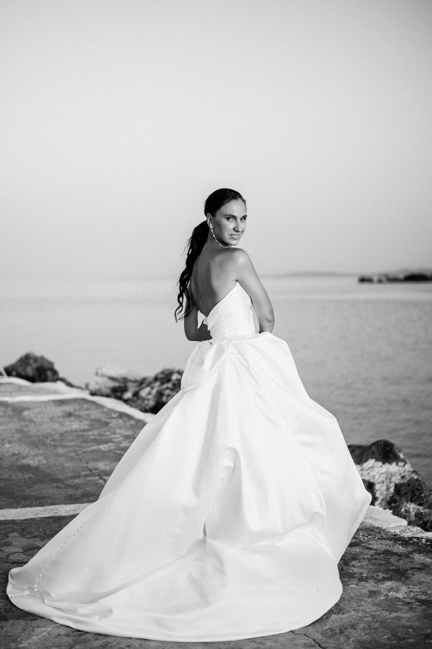 Black and white bridal portrait showcasing flowing white gown against coastal landscape.
