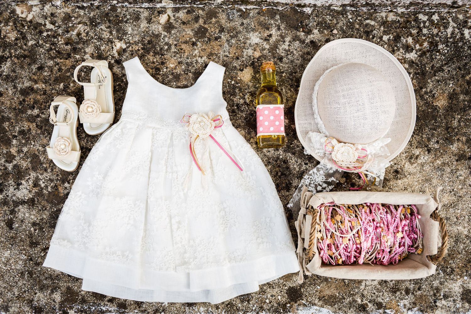 White baby dress, sun hat, and accessories laid out on stone surface with pink details.