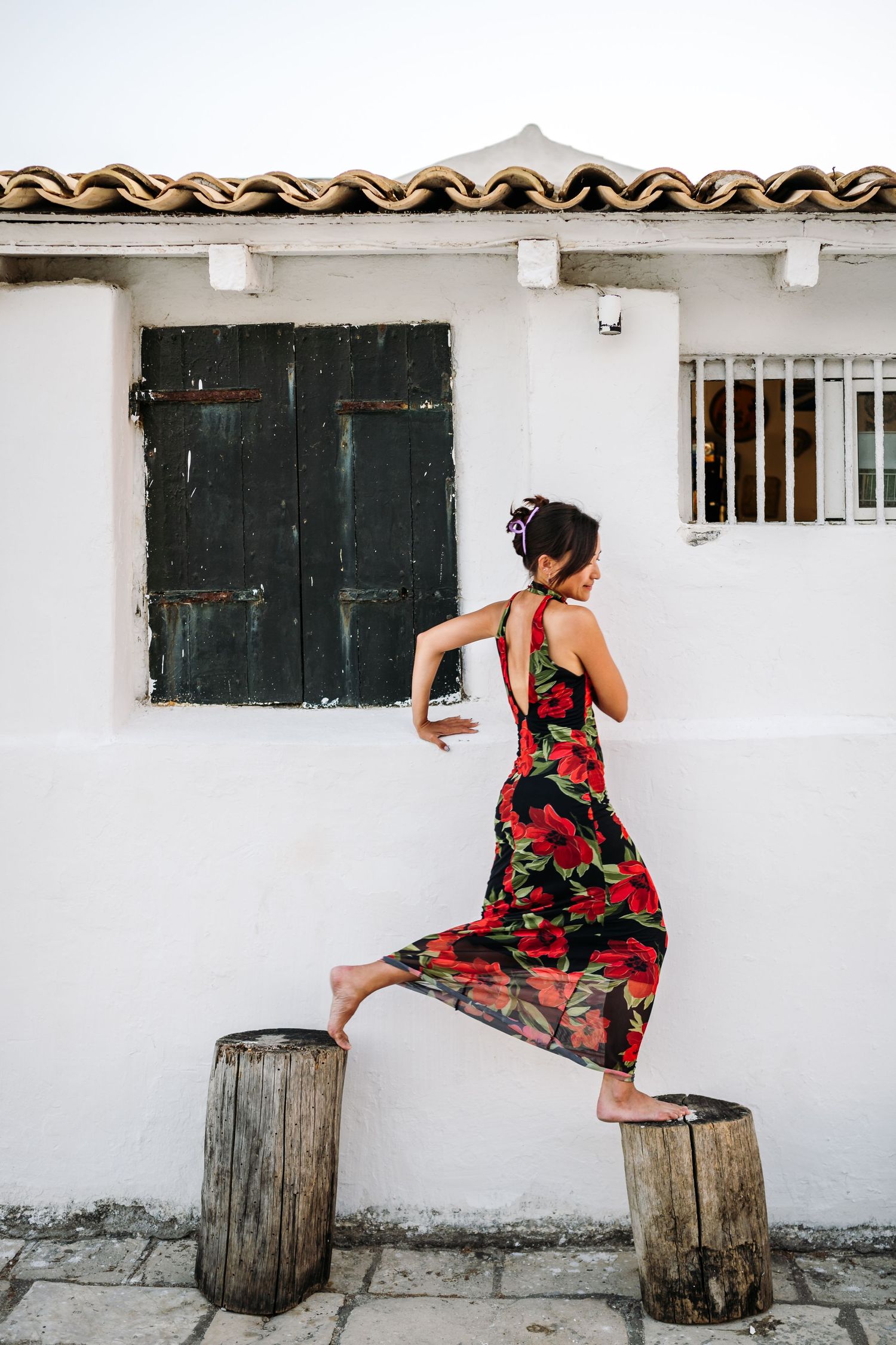 Woman in floral dress sitting on wooden barrels against white building wall.