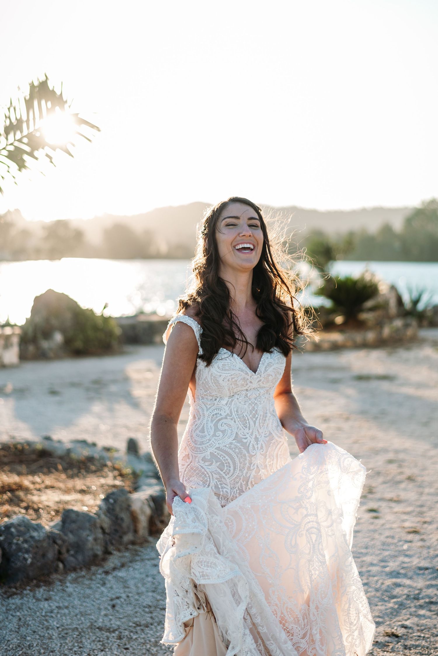 Bride in lace wedding dress laughs joyfully on sandy beach at sunset.