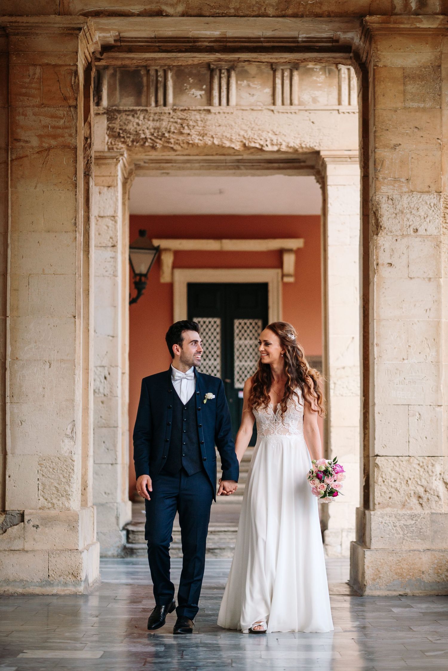 Elegant couple walking through historic stone colonnade during wedding photo session.