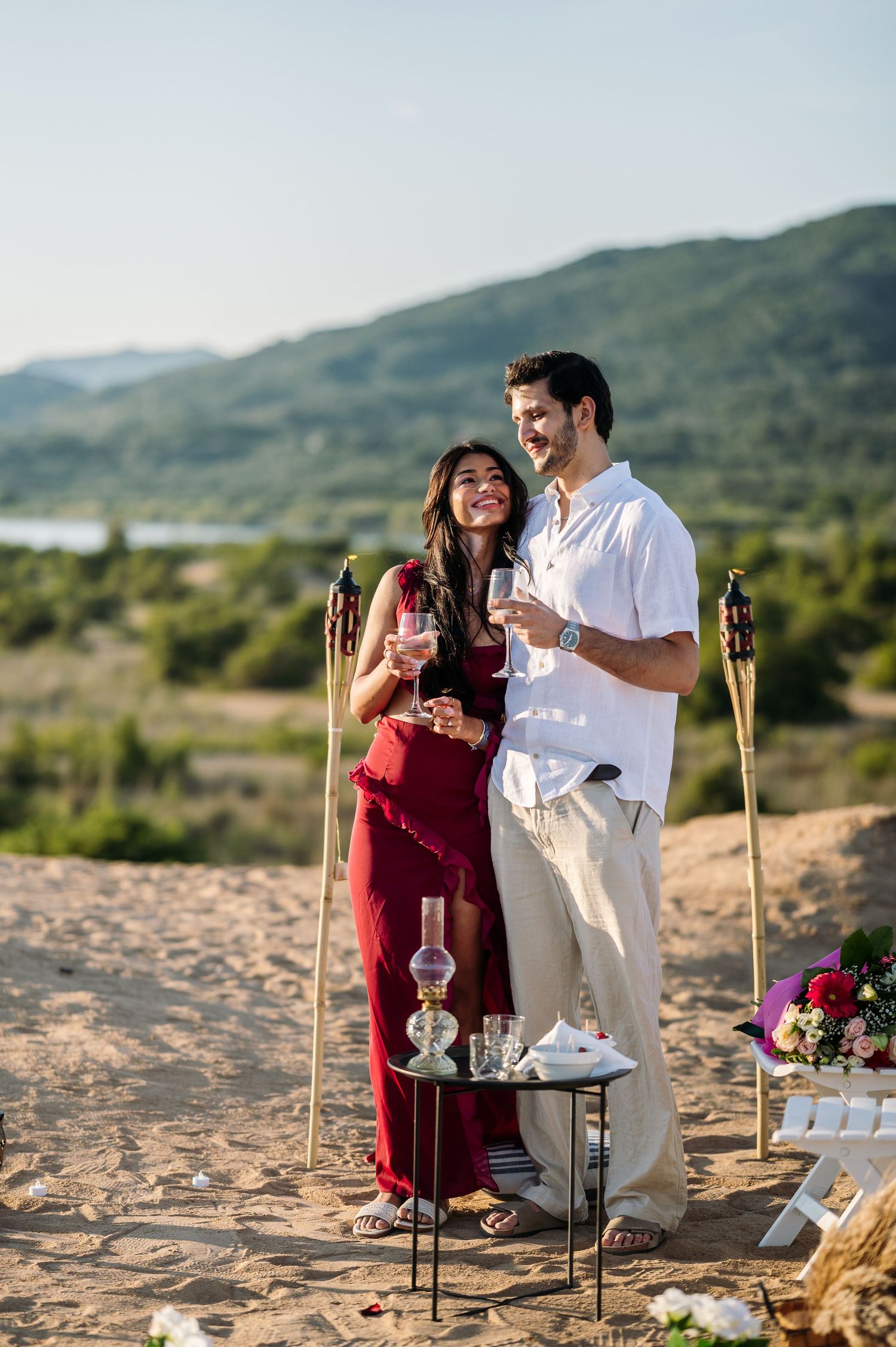 A couple celebrating their engagement at a decorated beach picnic setup with tiki torches.