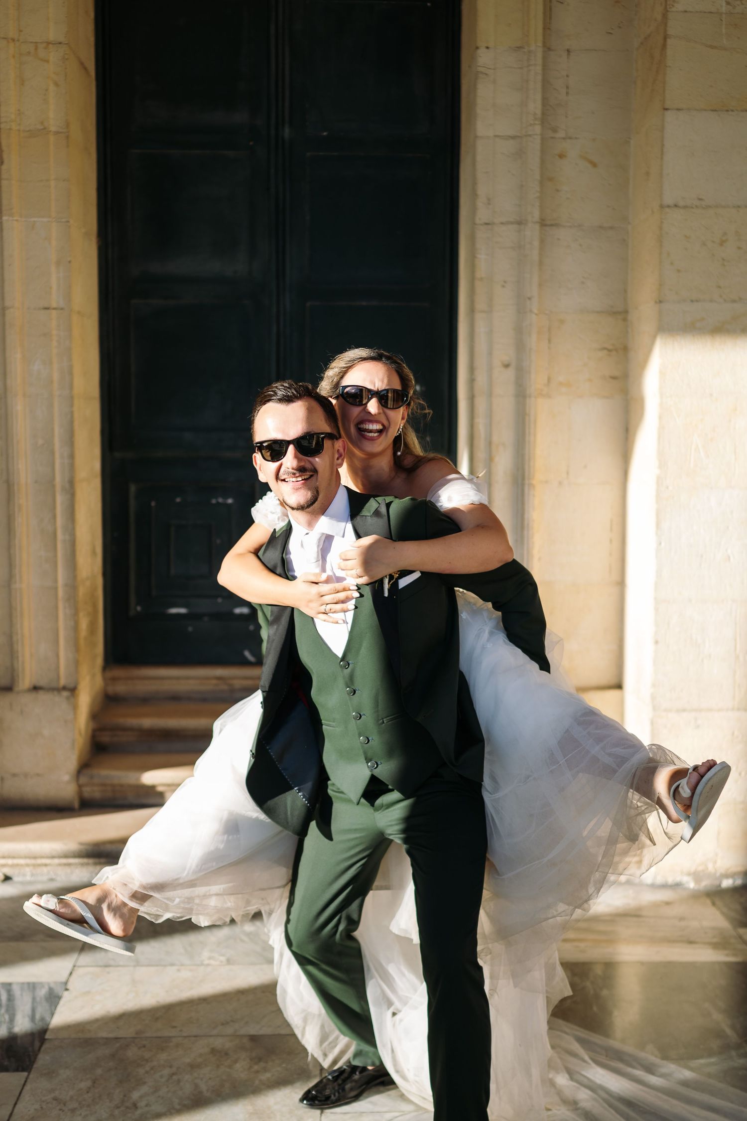 Playful wedding couple in sunglasses and formal attire pose together in an elegant outdoor colonnade.