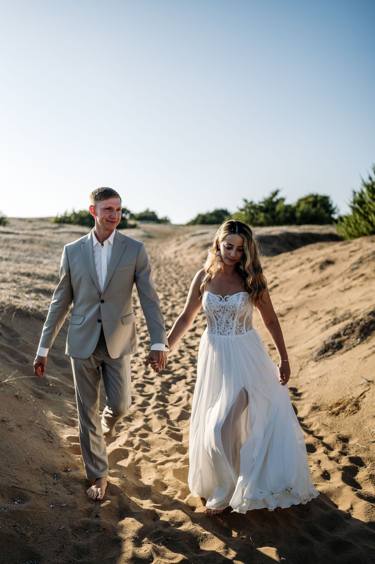 A couple walks hand in hand along a sandy beach path wearing elegant wedding attire.