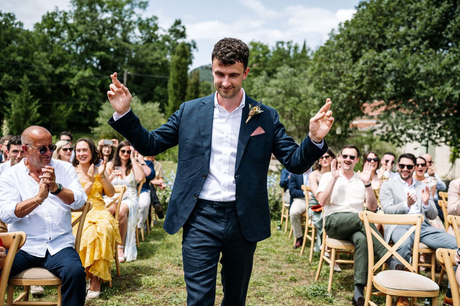 Wedding guests seated in wooden chairs at an outdoor ceremony surrounded by greenery.