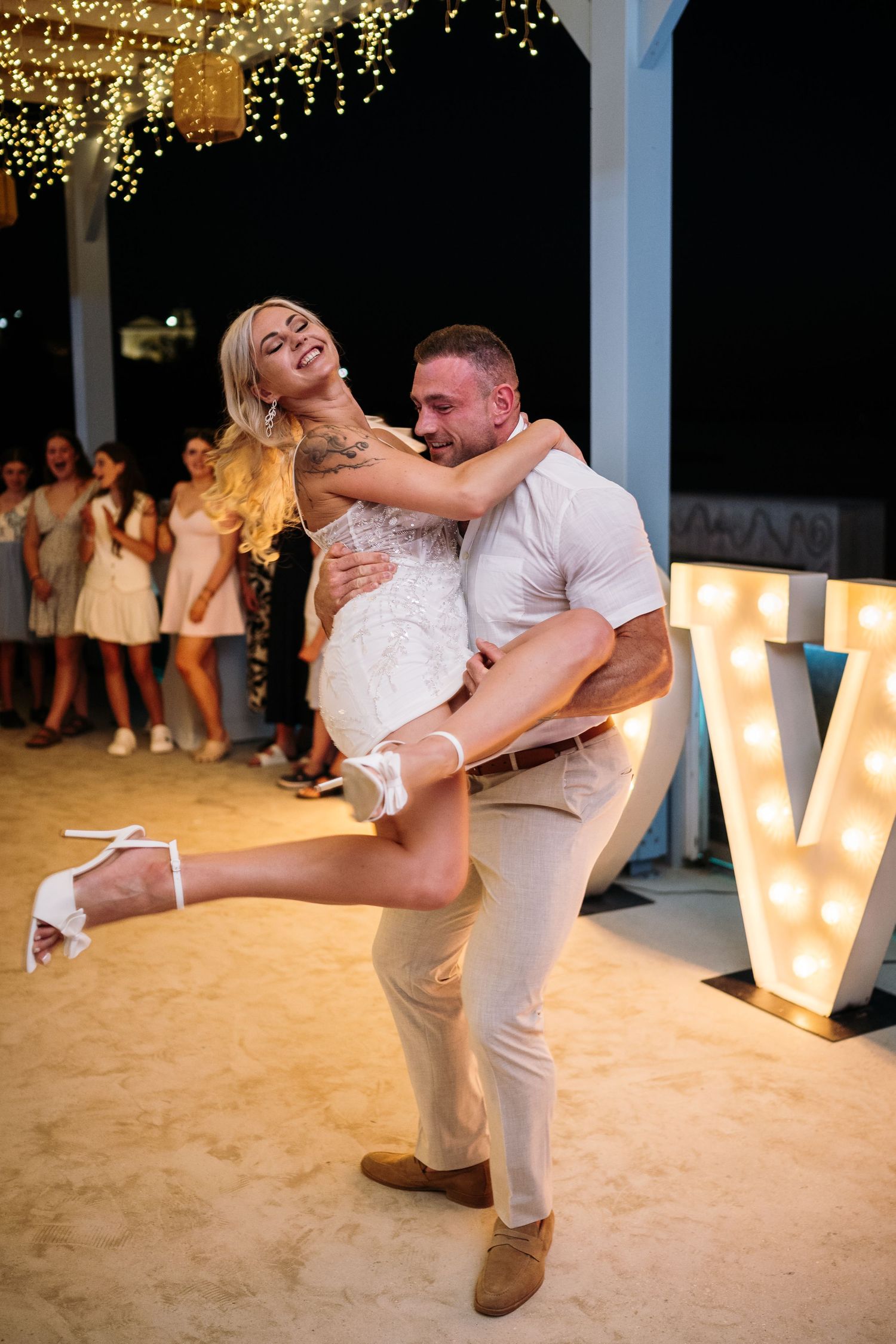 Dancing couple performs dip move under string lights at beach wedding.