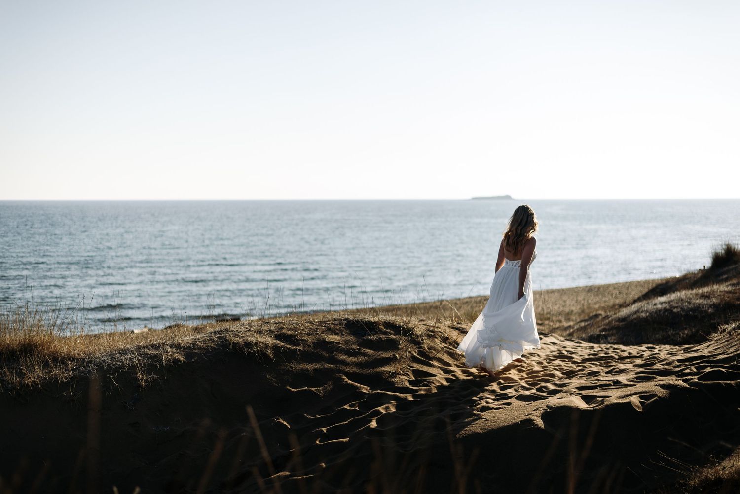 A bride in a flowing white dress stands alone on a sandy cliff overlooking the sea.