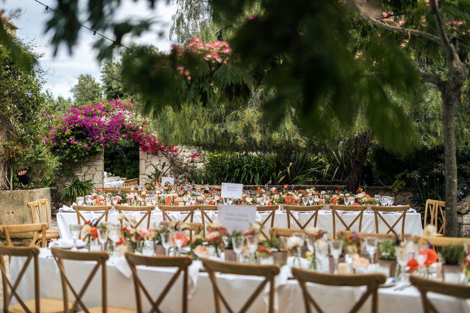 Elegant outdoor wedding reception setup with wooden cross-back chairs and long tables under trees.