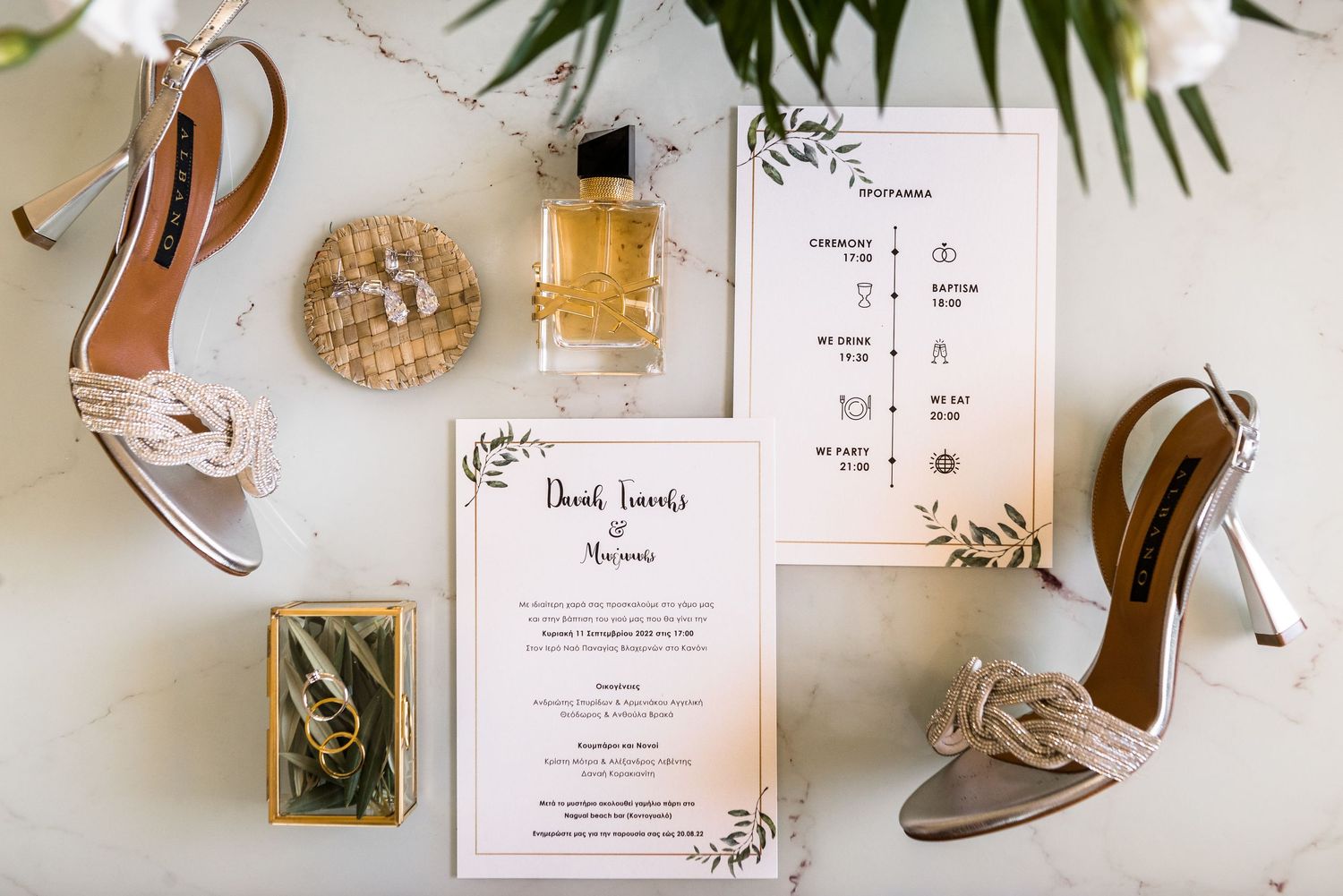 Wedding details flatlay with invitation cards, rings, and high heel shoes on white background.