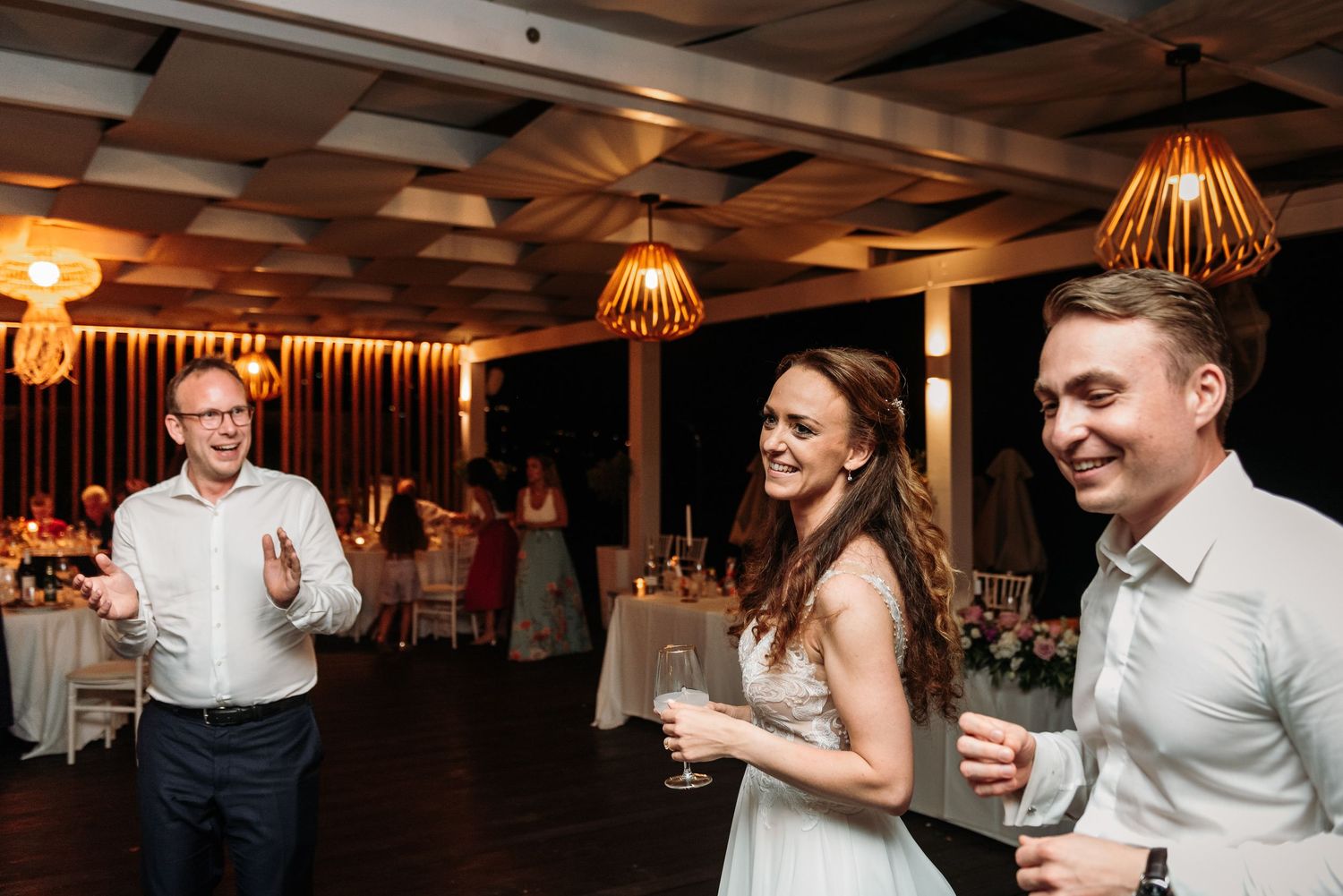 Wedding reception guests dancing and celebrating on a dimly lit dance floor with warm pendant lighting.