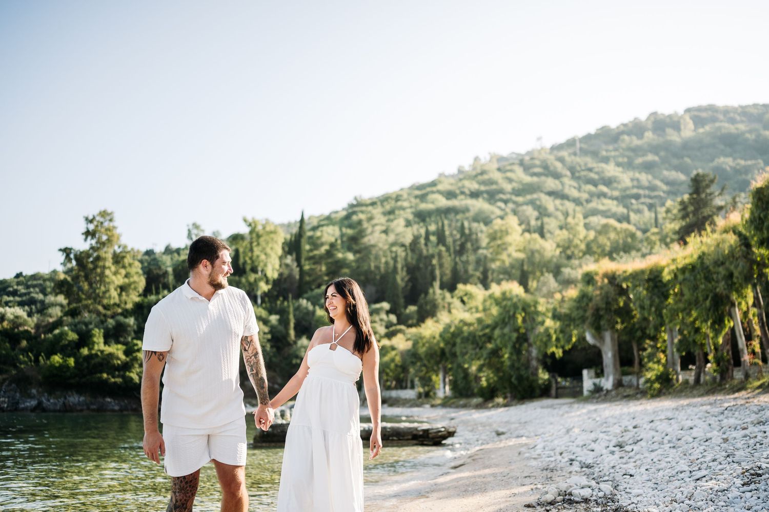 A couple strolling along a picturesque Mediterranean beach with lush green hills in the background.