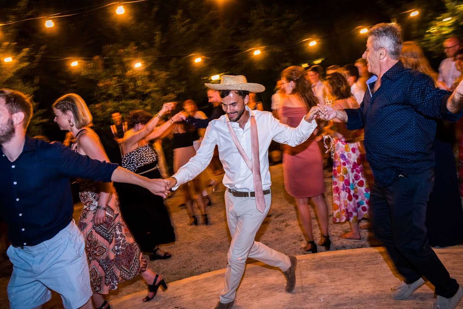 Lively group dancing outdoors under festive string lights at a wedding celebration.