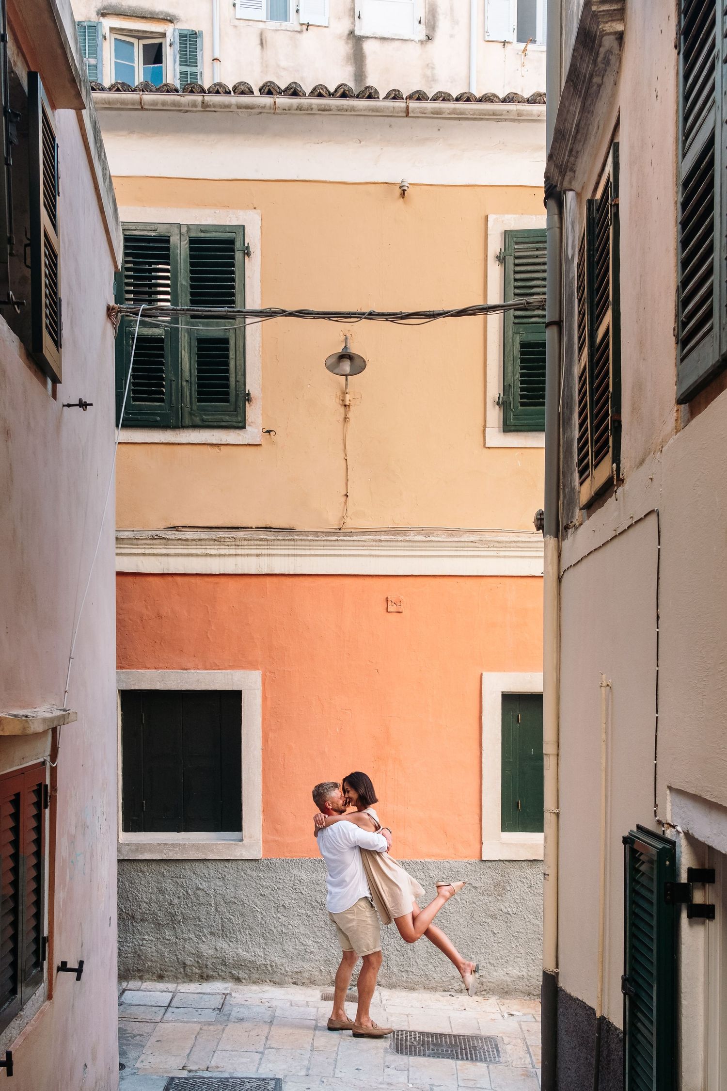 Romantic couple shares playful moments in a Mediterranean alleyway with peach-colored walls and green shutters.