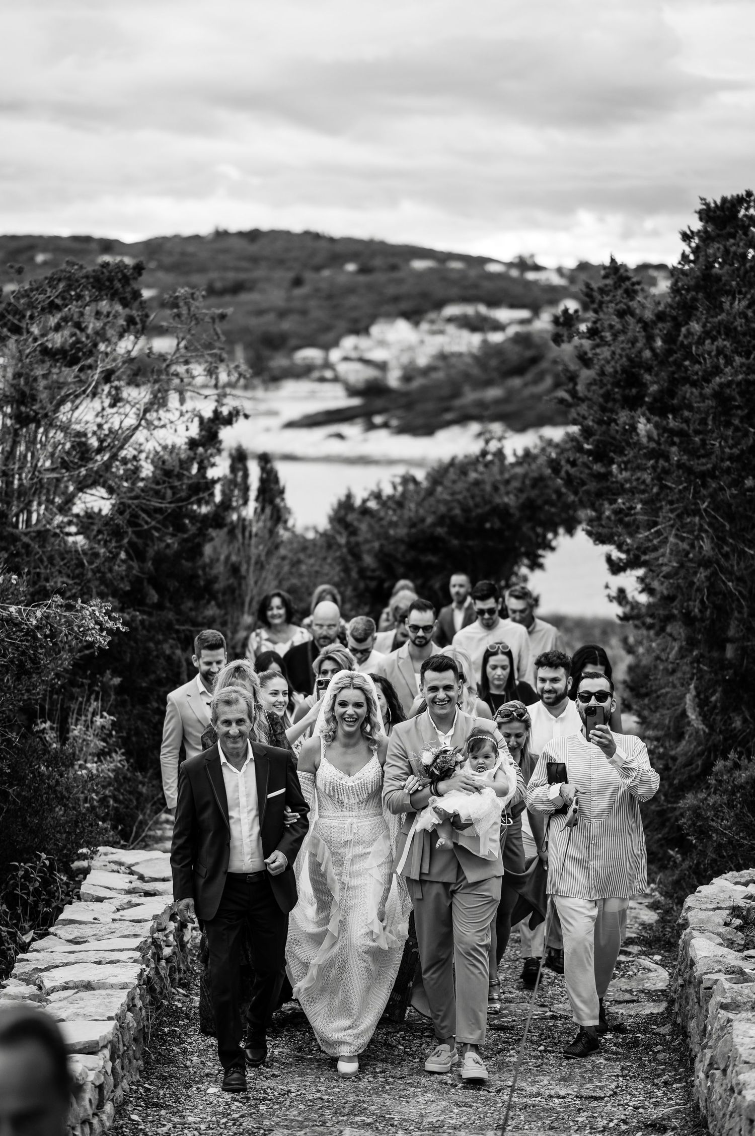 Wedding party and guests walk along a scenic hillside path surrounded by lush greenery.