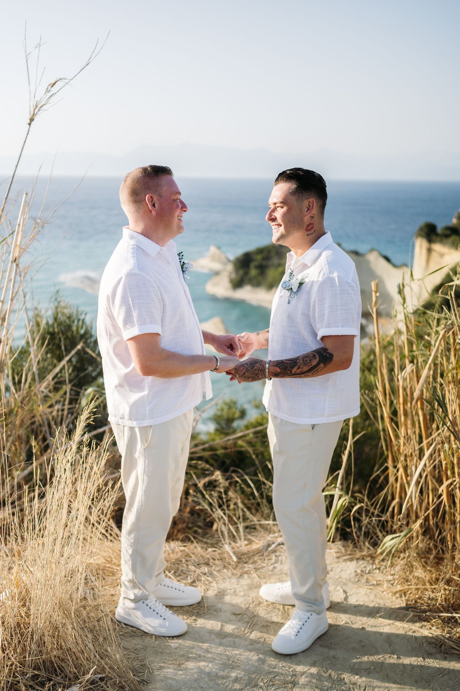 Couple in coordinated white outfits sharing intimate moment on clifftop overlooking ocean.