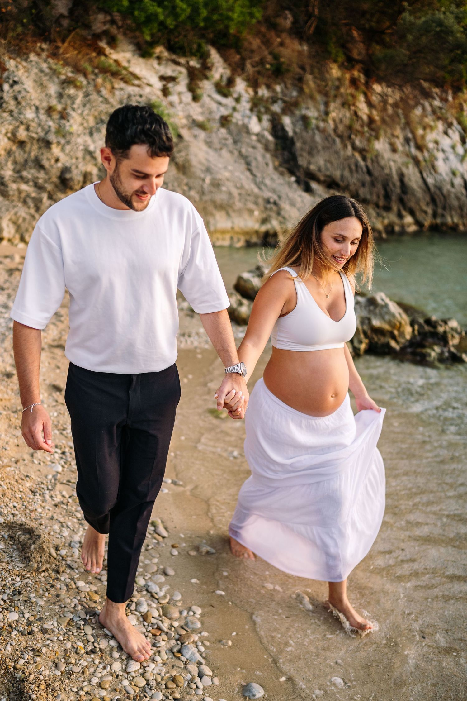 Couple playfully walks along rocky beach shoreline in white summer attire.