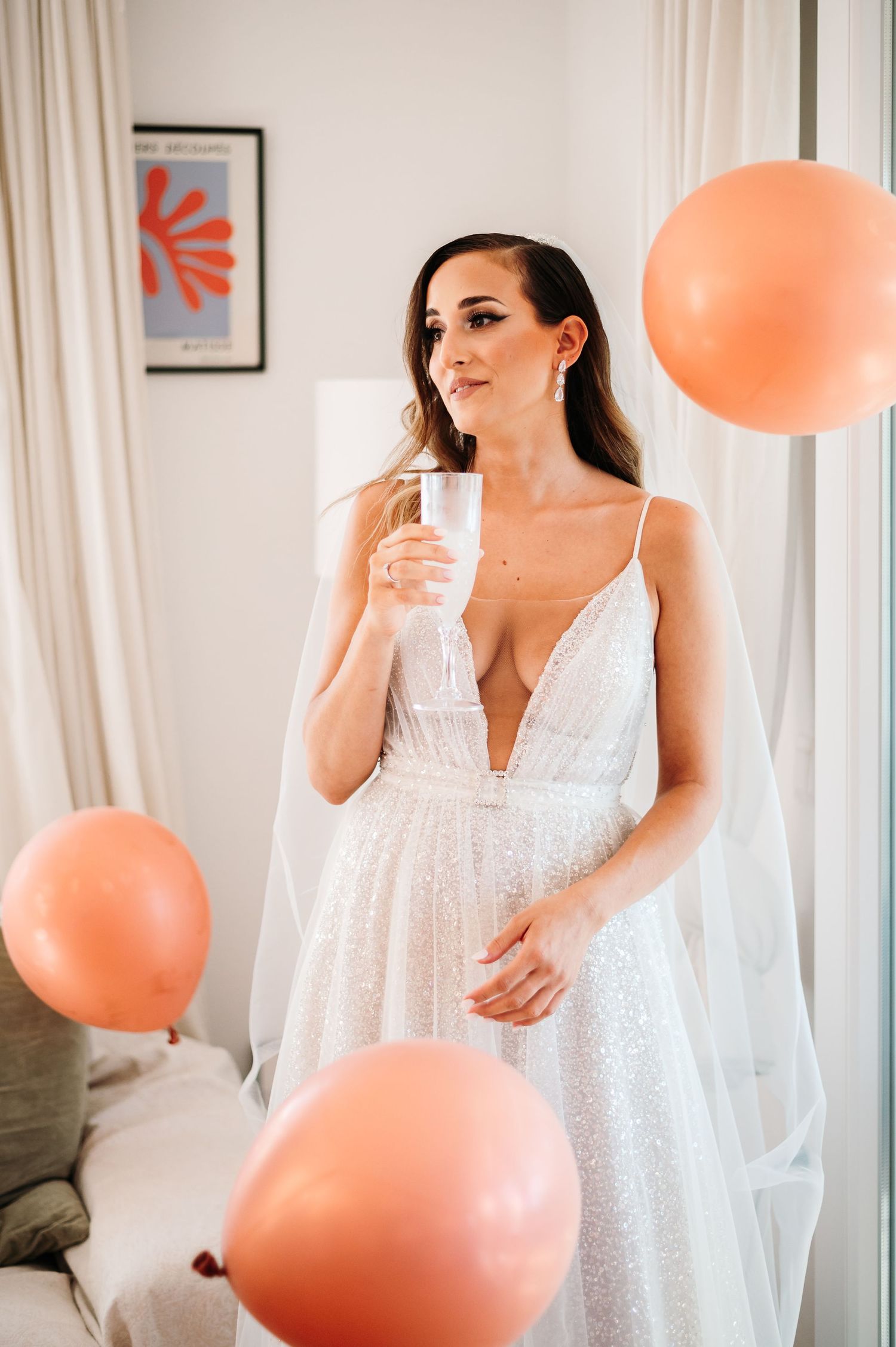 A white lace dress hangs in a bright room with coral colored balloons floating nearby.