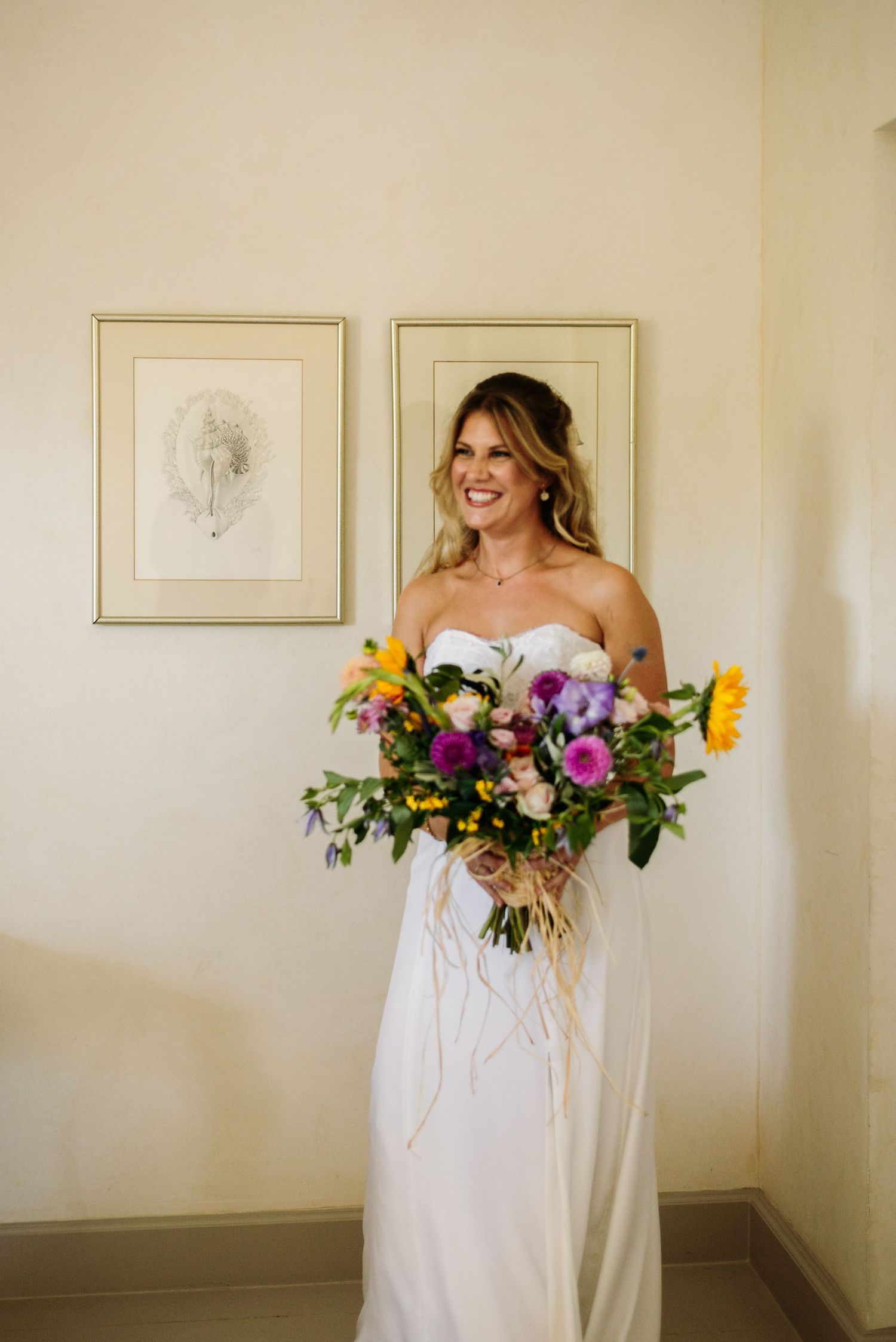 Elegant white dress with vibrant wildflower bouquet against cream-colored wall with framed artwork.