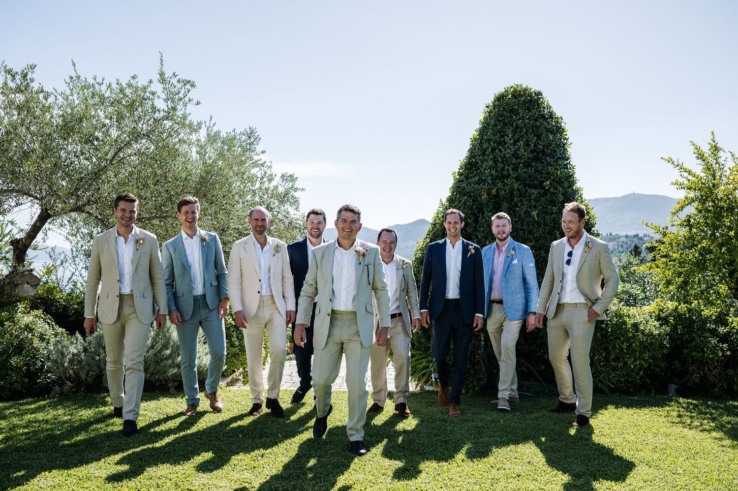 Wedding party in light colored suits posed outdoors against mountain backdrop.