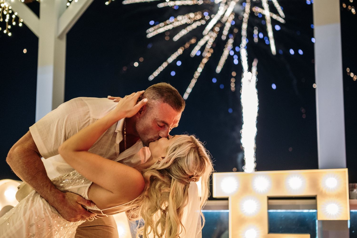 Couple dancing with fireworks display in background at beach wedding.