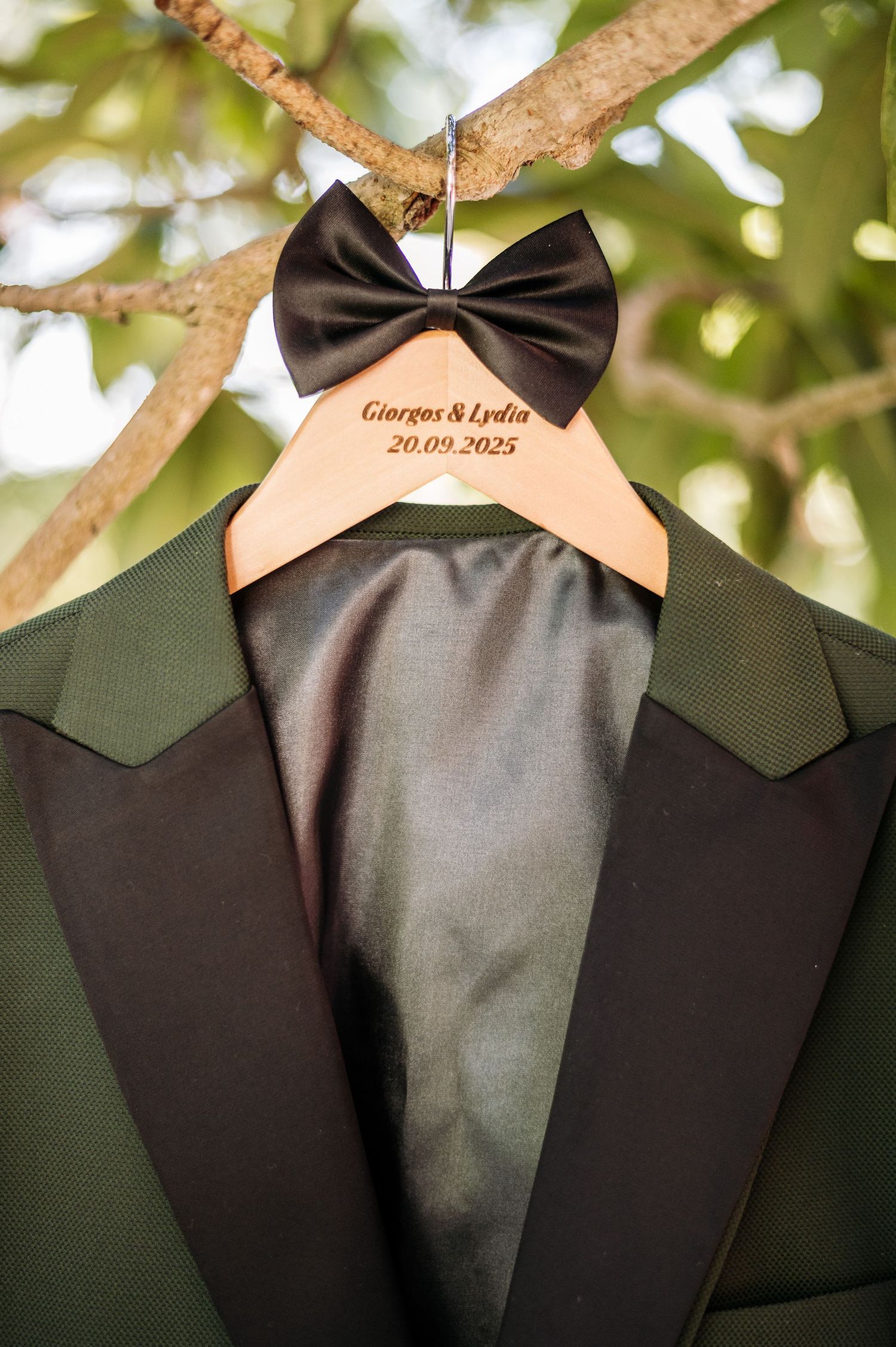 Wedding suit jacket and bowtie hanging on wooden hanger against green foliage background.