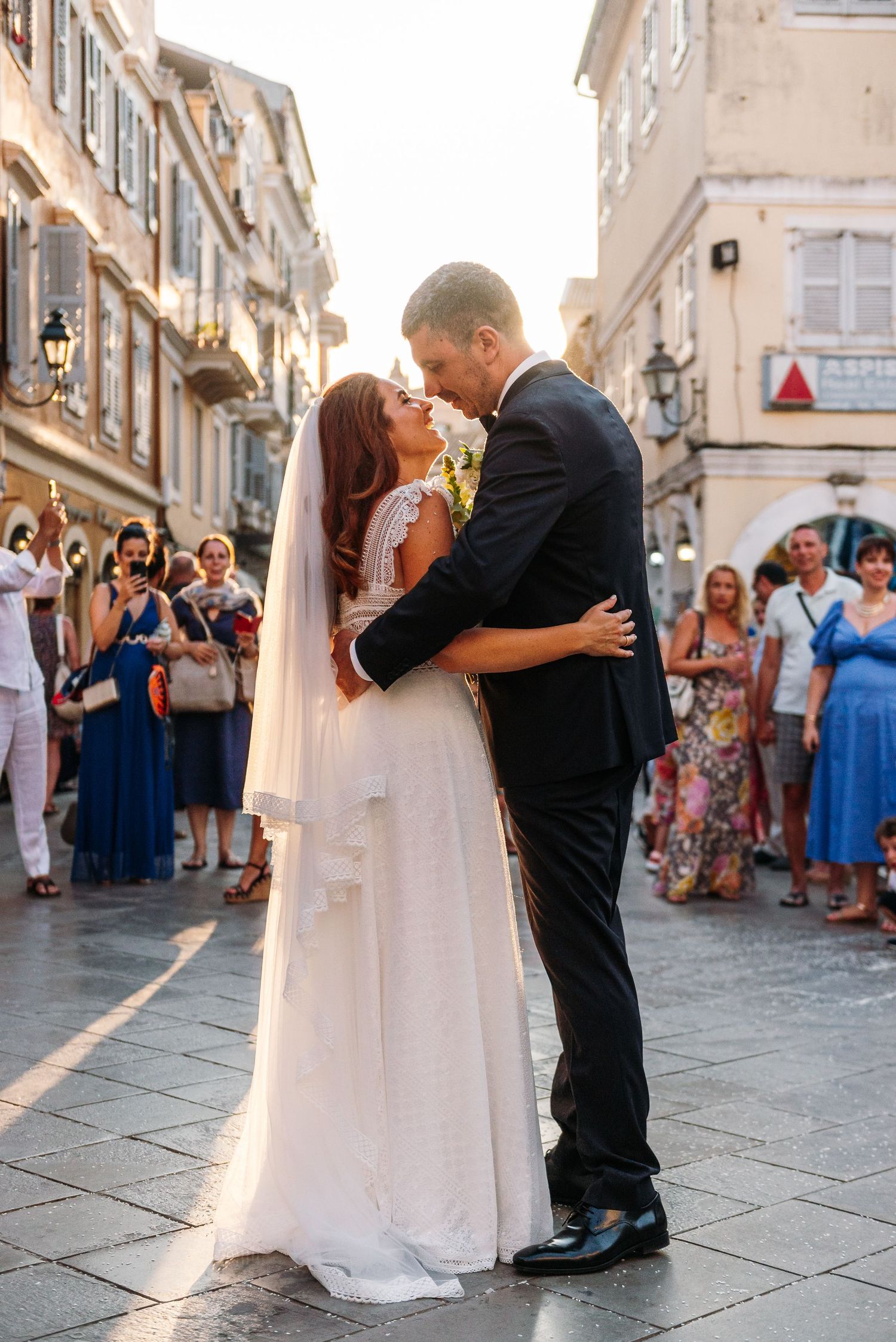 A romantic wedding moment in a European city street with guests in blue dresses watching in the background.