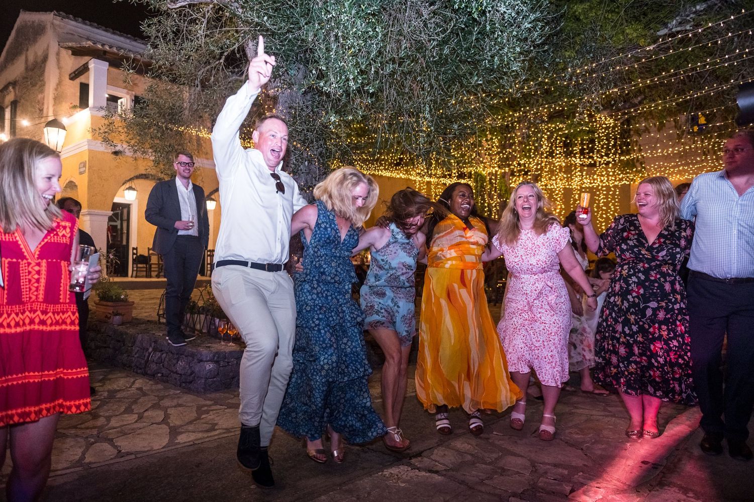 Wedding guests dancing energetically under canopy of string lights at outdoor reception.