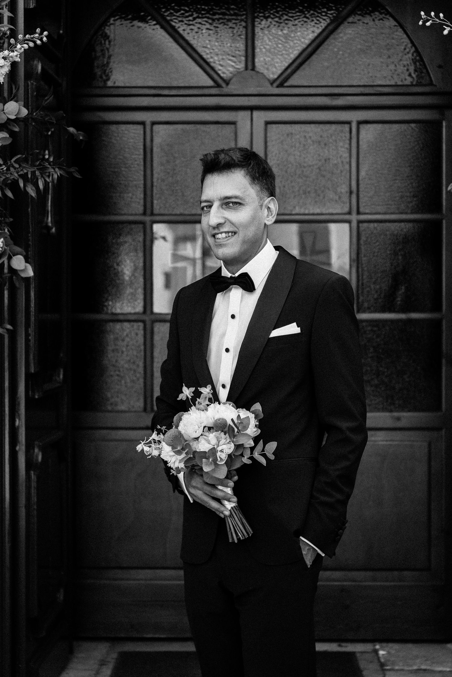 Black and white portrait of groomsman holding wedding bouquet against wooden paneling.