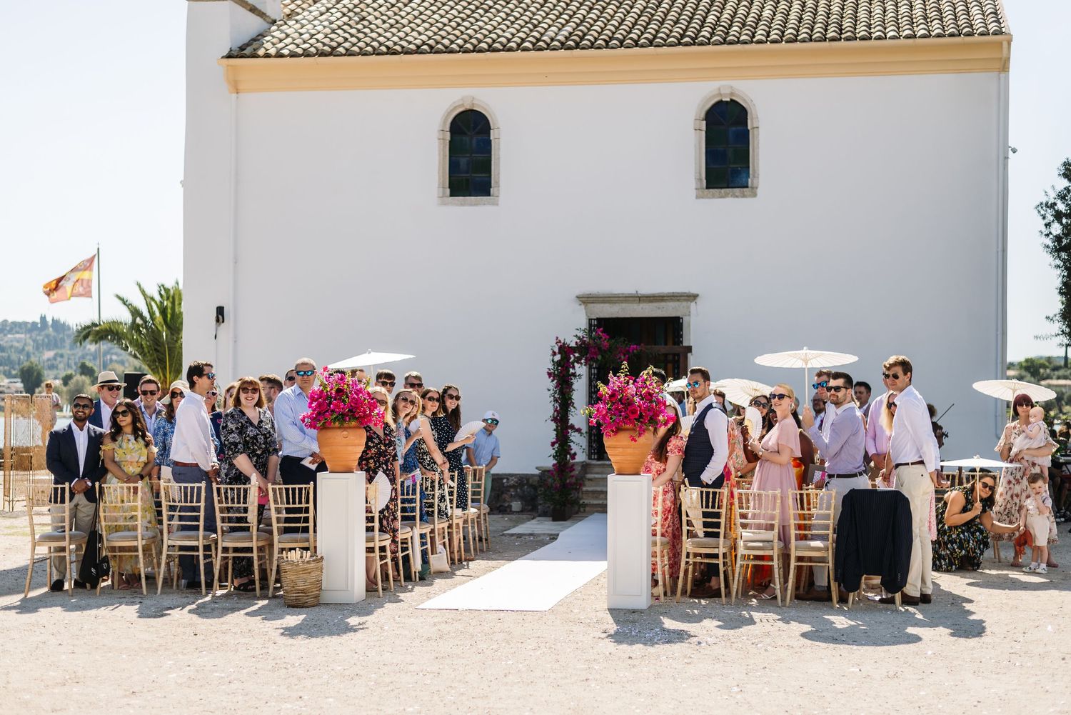 Wedding guests seated outside white Mediterranean style building for ceremony.