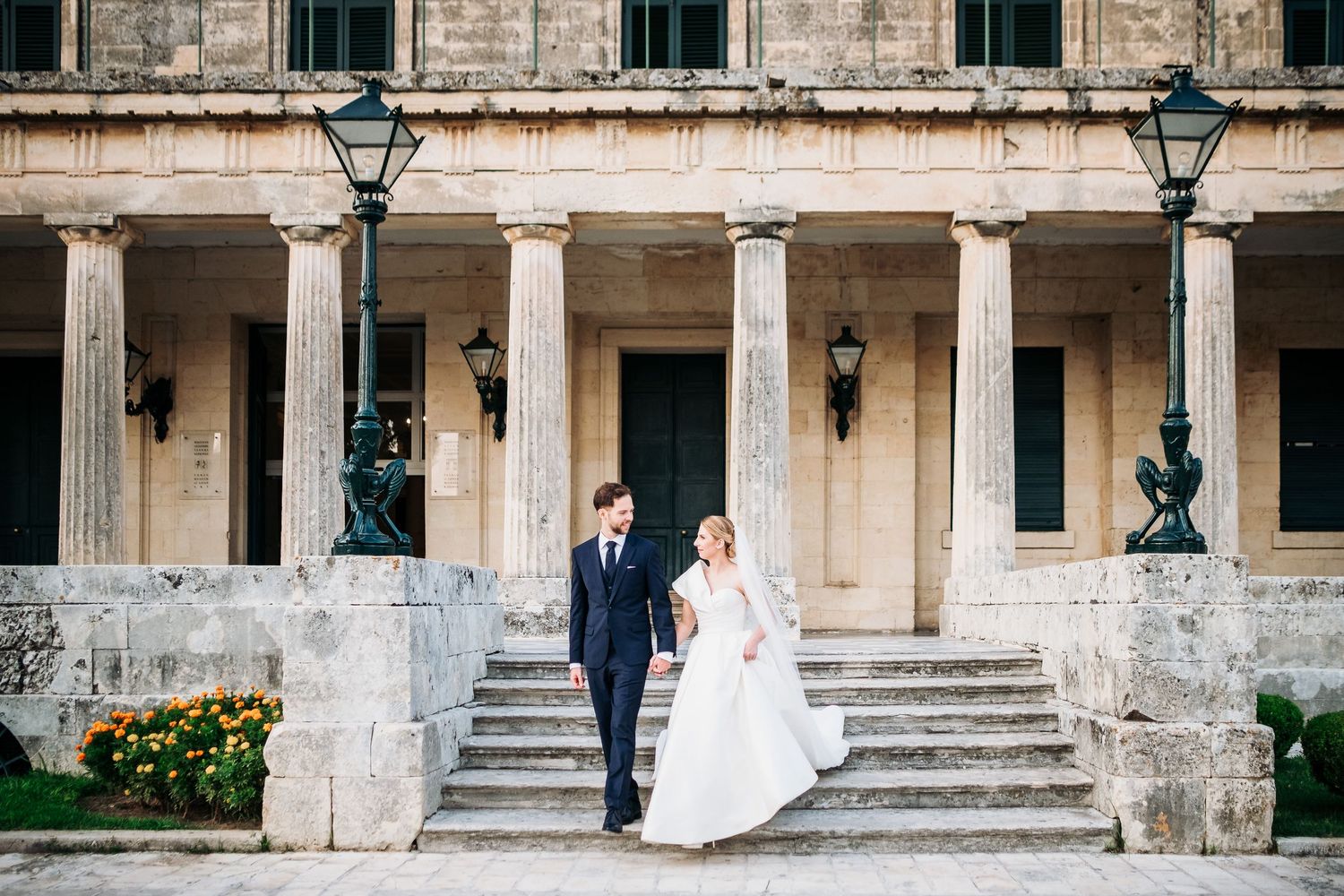 Wedding couple and guests pose along a grand stone colonnade exterior.