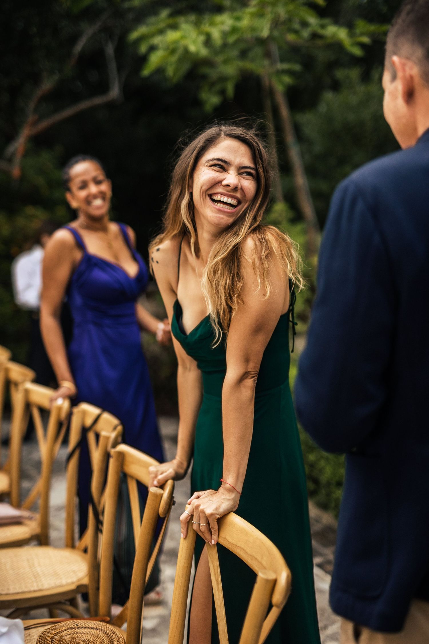 Wedding guests in elegant attire smile and laugh while seated in wooden chairs at an outdoor venue.