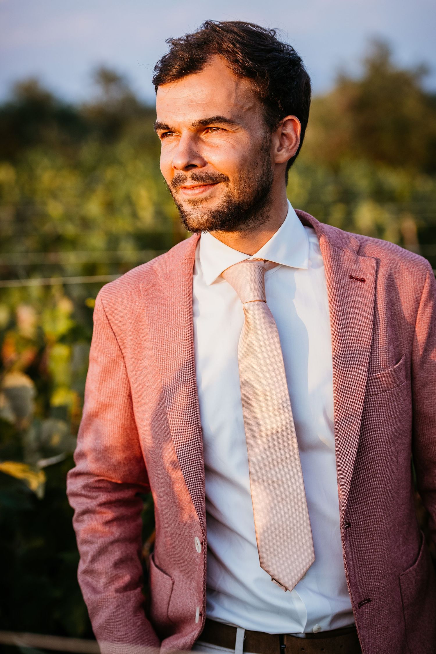 Groom in dusty rose suit jacket and white tie smiling in warm sunlight.