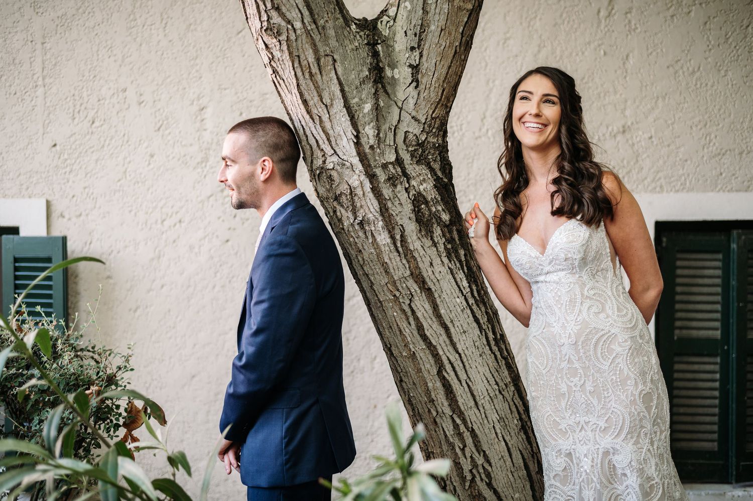 A couple in wedding attire poses playfully on opposite sides of a tree trunk during their first look photo session.