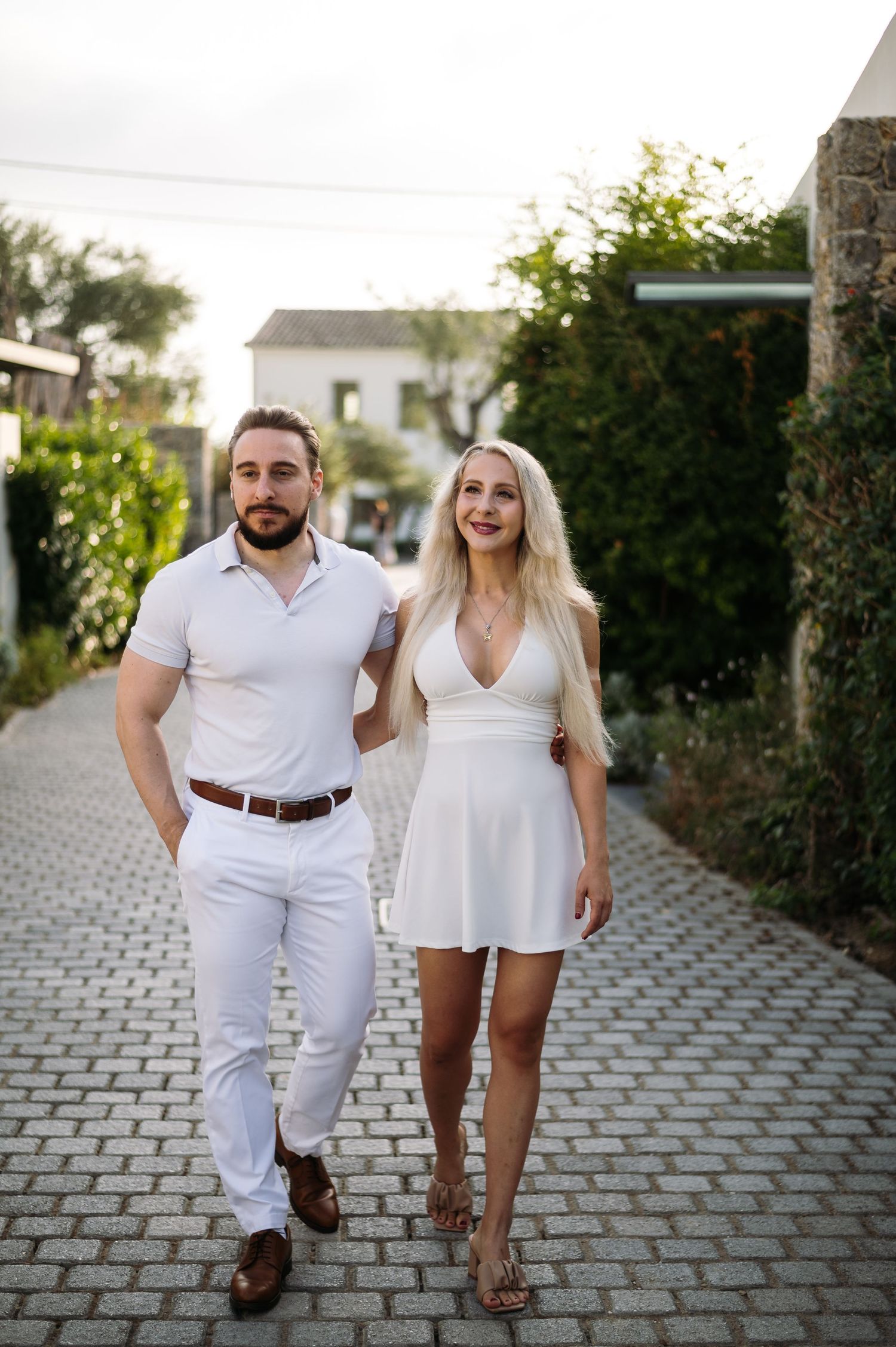 A couple wearing coordinated white outfits walks together on a stone pathway.