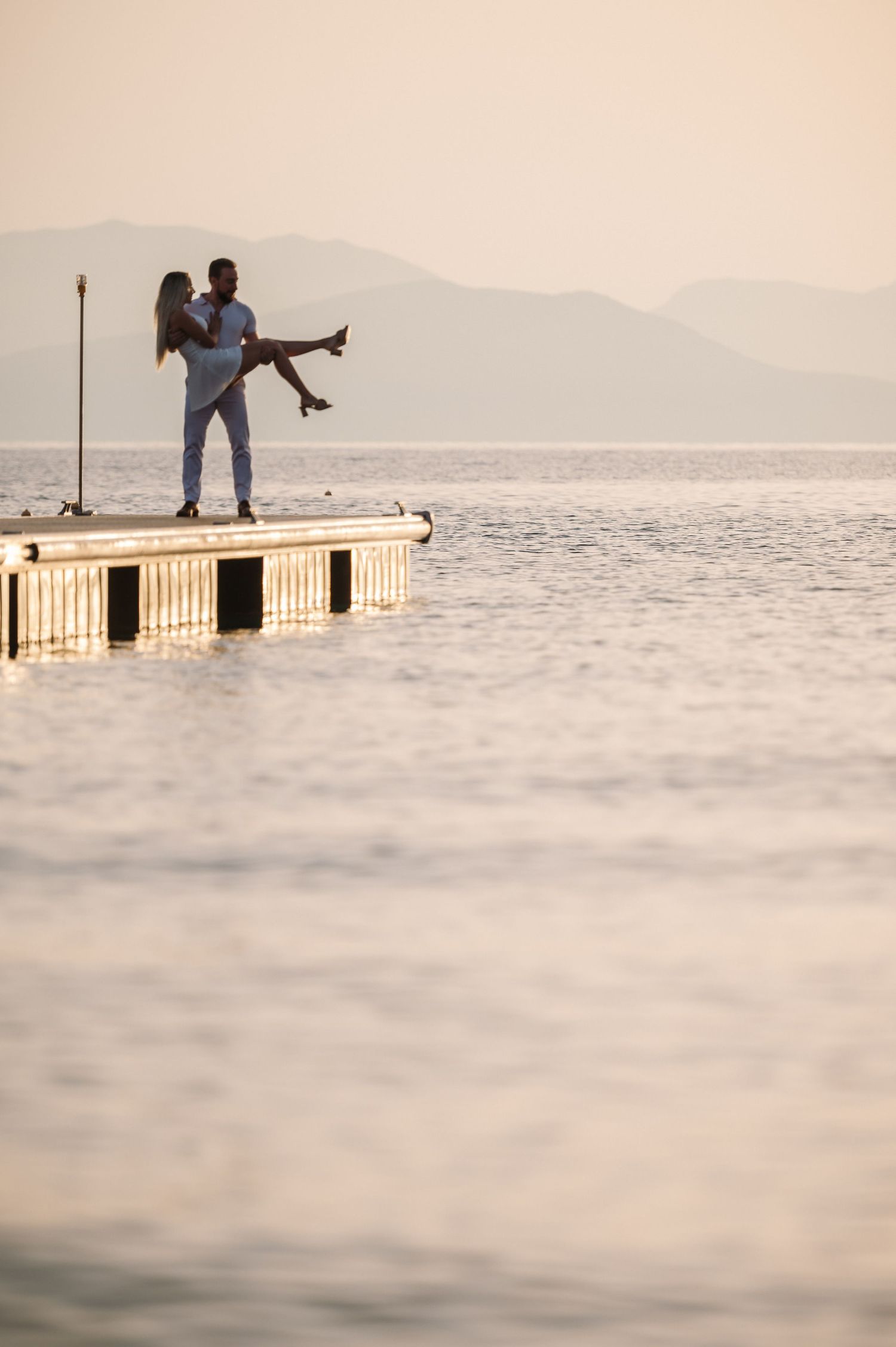 Silhouettes of couple on wooden pier during sunset with mountains in background.